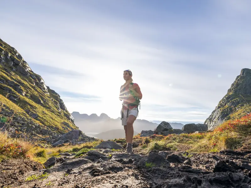 Wanderin auf einem steinigen Pfad in einem Tal der Lofoten, im Hintergrund die von der Sonne beschienene Fjordlandschaft.