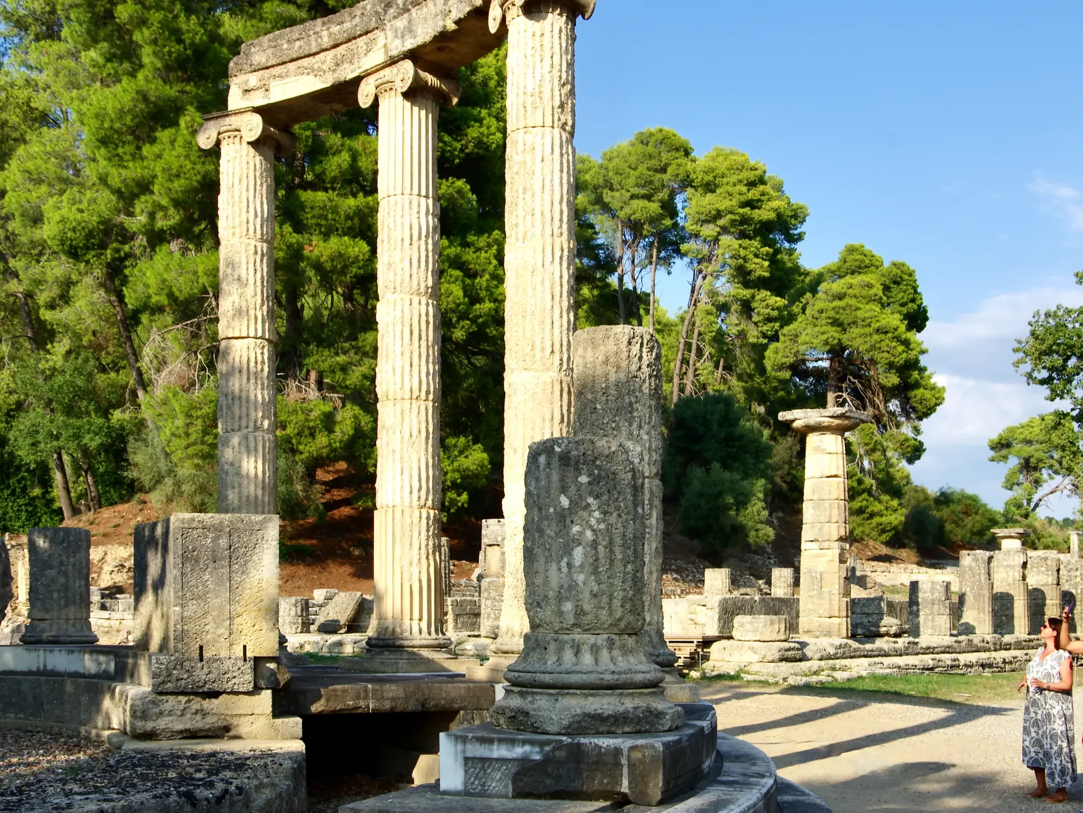 Zwei Frauen auf Studienreise betrachten die sonnenbeschienenen Säulen des Philippeion der antiken Ausgrabungen von Olympia auf dem Peloponnes.