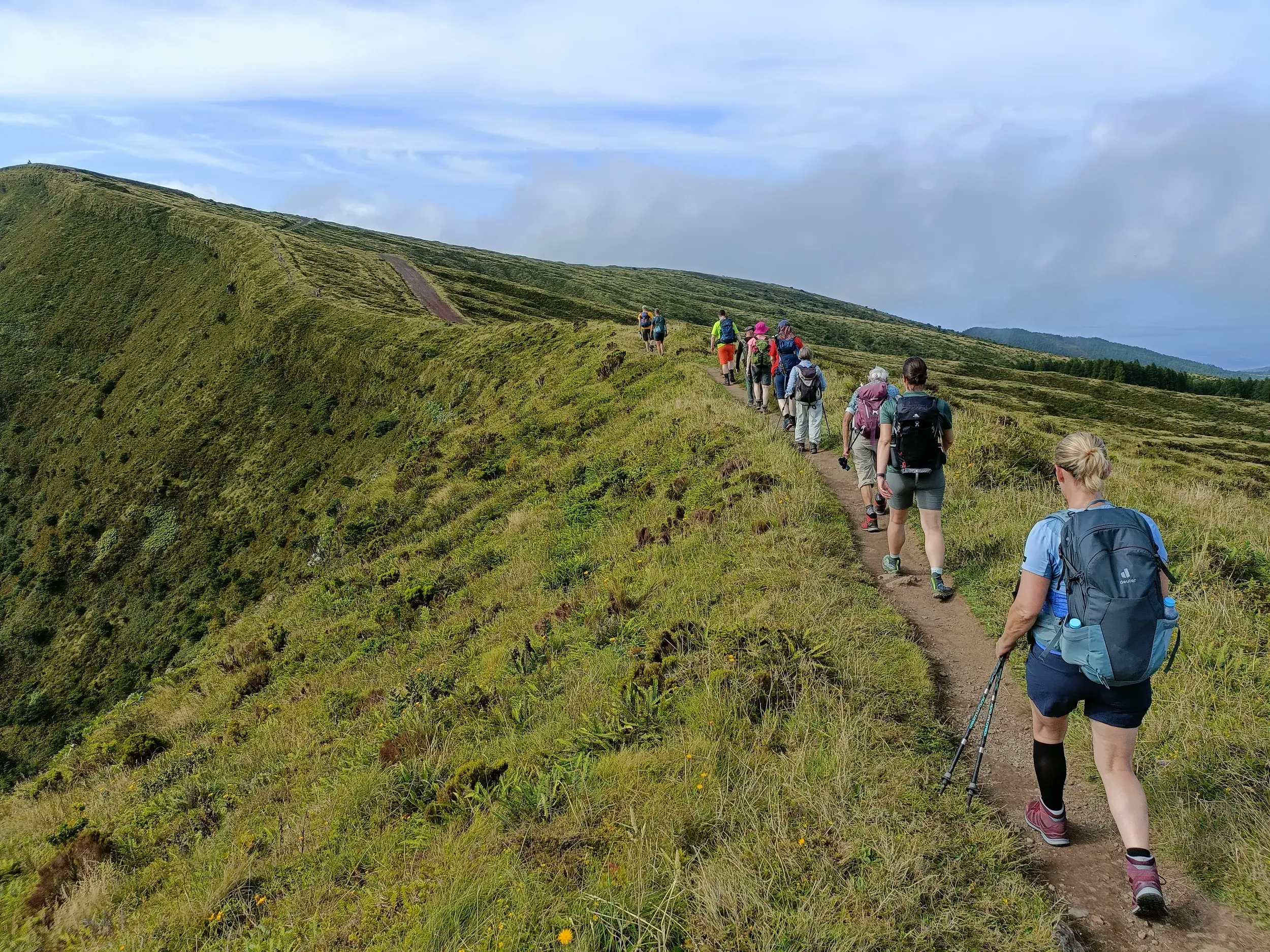 Hauser-Wandergruppe beim Trekking auf einem schmalen Pfad entlang eines grünen, grasbewachsenen Kraterrands auf den Azoren.