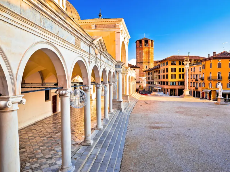 Blick vom Arkadengang der Loggia di San Giovanni auf die sonnige Piazza della Libertà in Udine mit historischen Gebäuden.