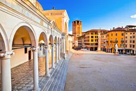 Blick vom Arkadengang der Loggia di San Giovanni auf die sonnige Piazza della Libertà in Udine mit historischen Gebäuden.