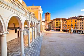 Blick vom Arkadengang der Loggia di San Giovanni auf die sonnige Piazza della Libertà in Udine mit historischen Gebäuden.