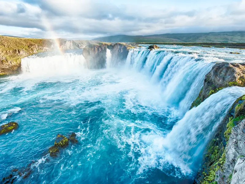Der hufeisenförmige Godafoss Wasserfall in Island, dessen türkise Wassermassen unter einem Regenbogen in die Tiefe stürzen.