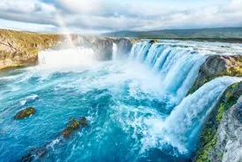 Der hufeisenförmige Godafoss Wasserfall in Island, dessen türkise Wassermassen unter einem Regenbogen in die Tiefe stürzen.