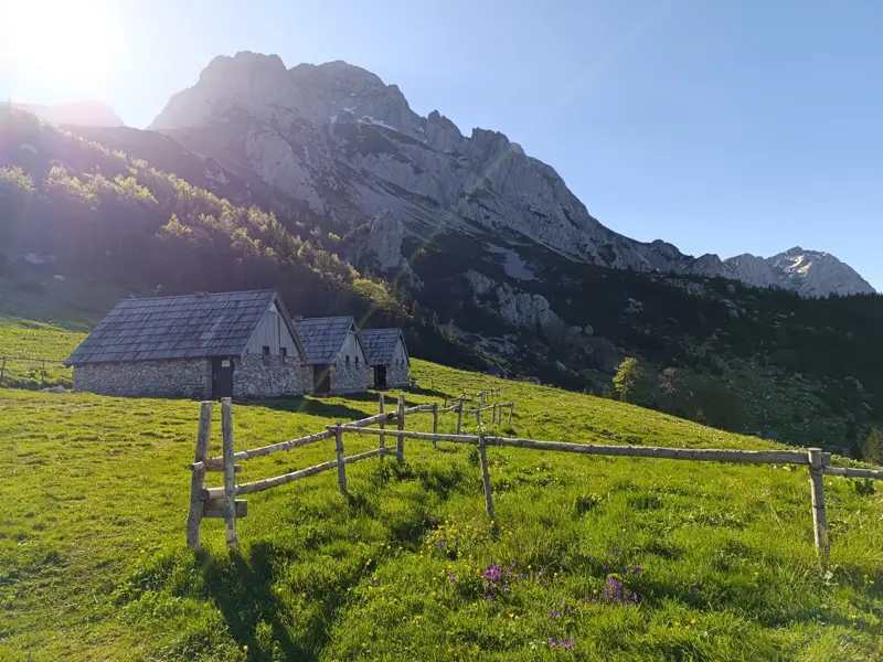 Drei rustikale Steinhütten auf einer saftig grünen Almwiese, im Hintergrund eine imposante Bergkette bei Sonnenaufgang.