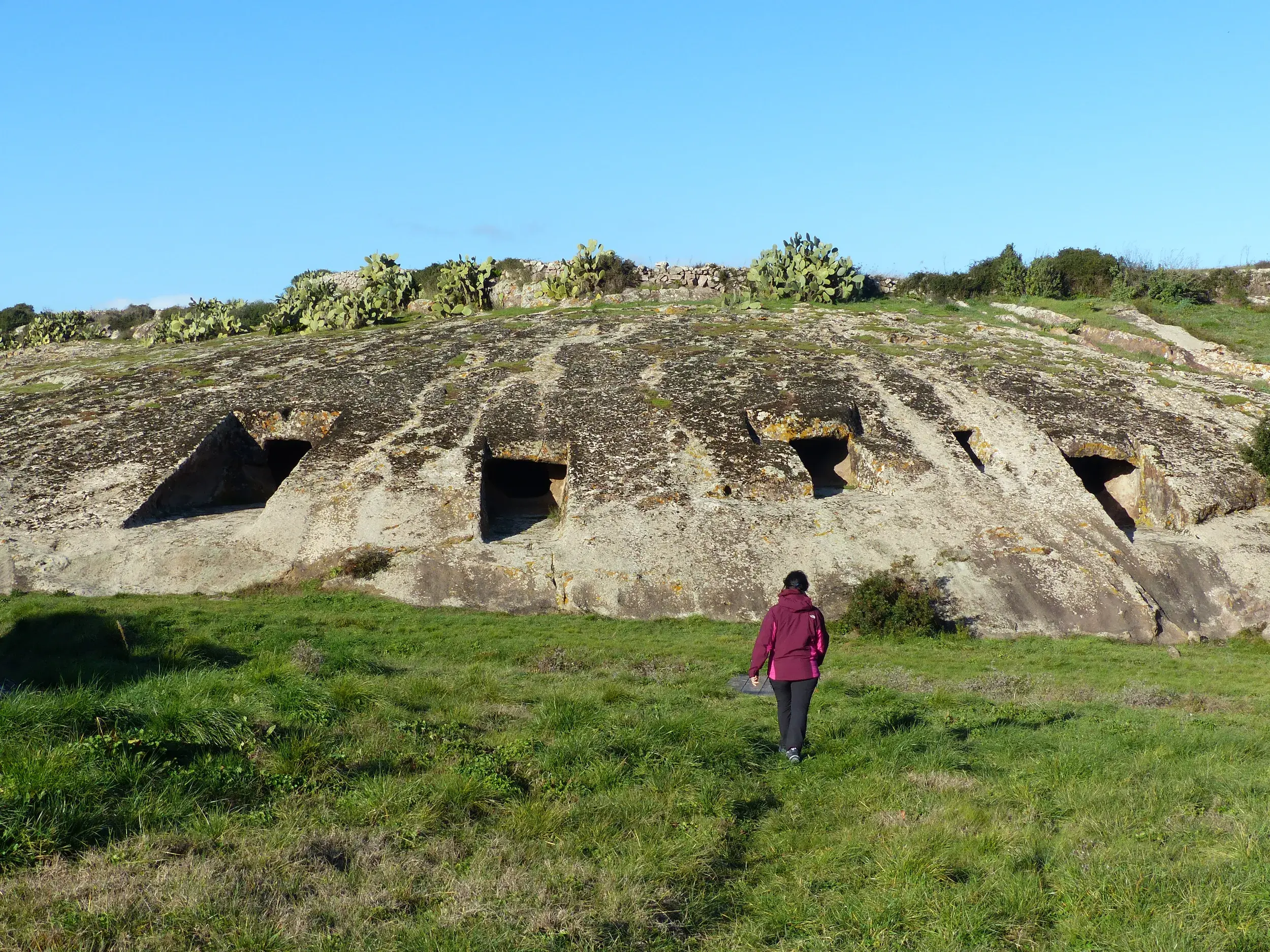 Eine Wanderin erkundet auf einer grünen Wiese die prähistorischen Höhlengräber Genna Salixi auf Sardinien bei klarem Himmel.