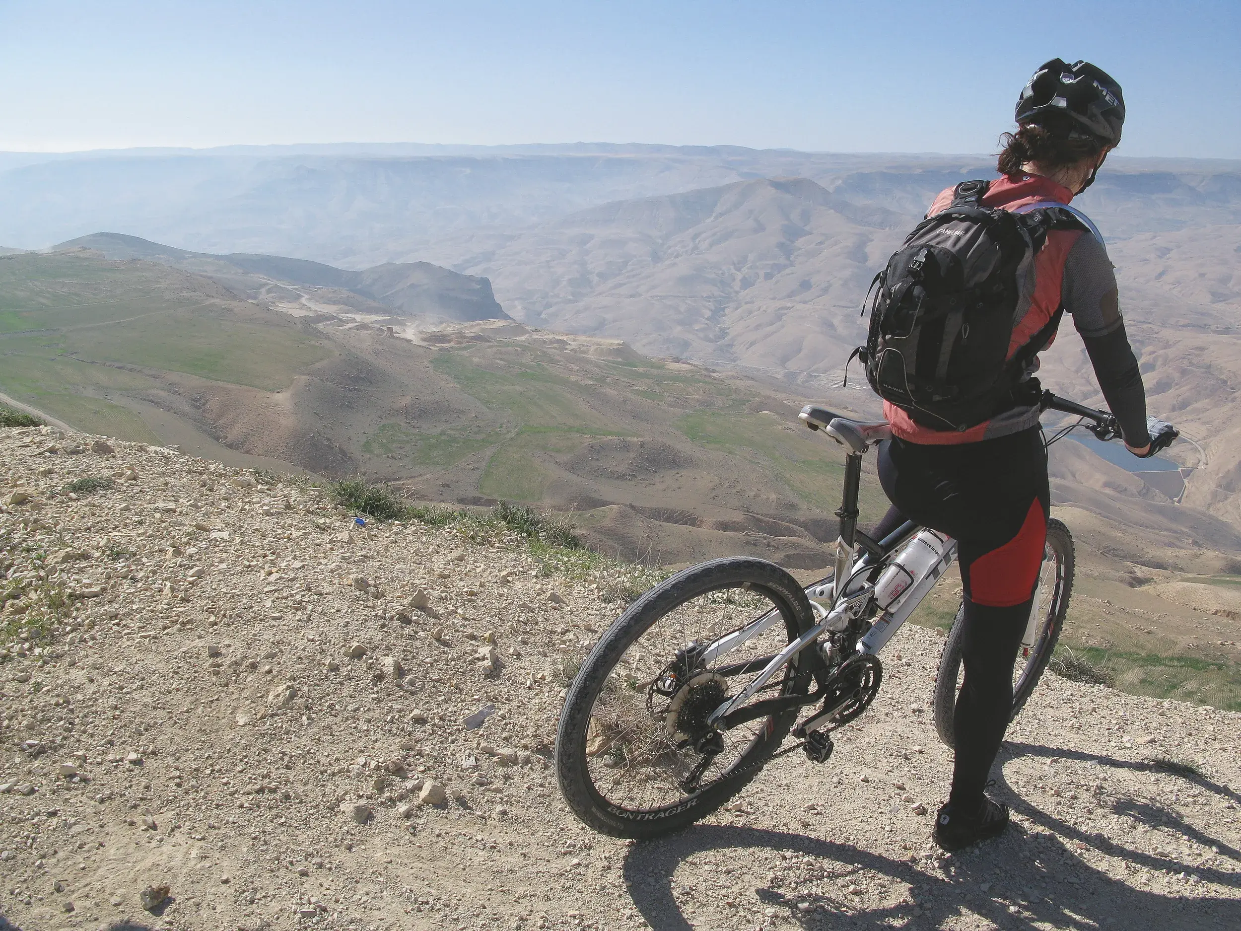 Mountainbikerin steht mit ihrem Rad auf einem Bergpfad und genießt den Panoramablick über die weite Wüstenlandschaft Jordaniens.