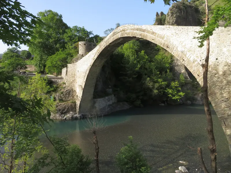 Imposante Steinbogenbrücke in der Zagori-Region, die sich bei Sonnenschein über einen Fluss inmitten von dichten grünen Wäldern spannt.