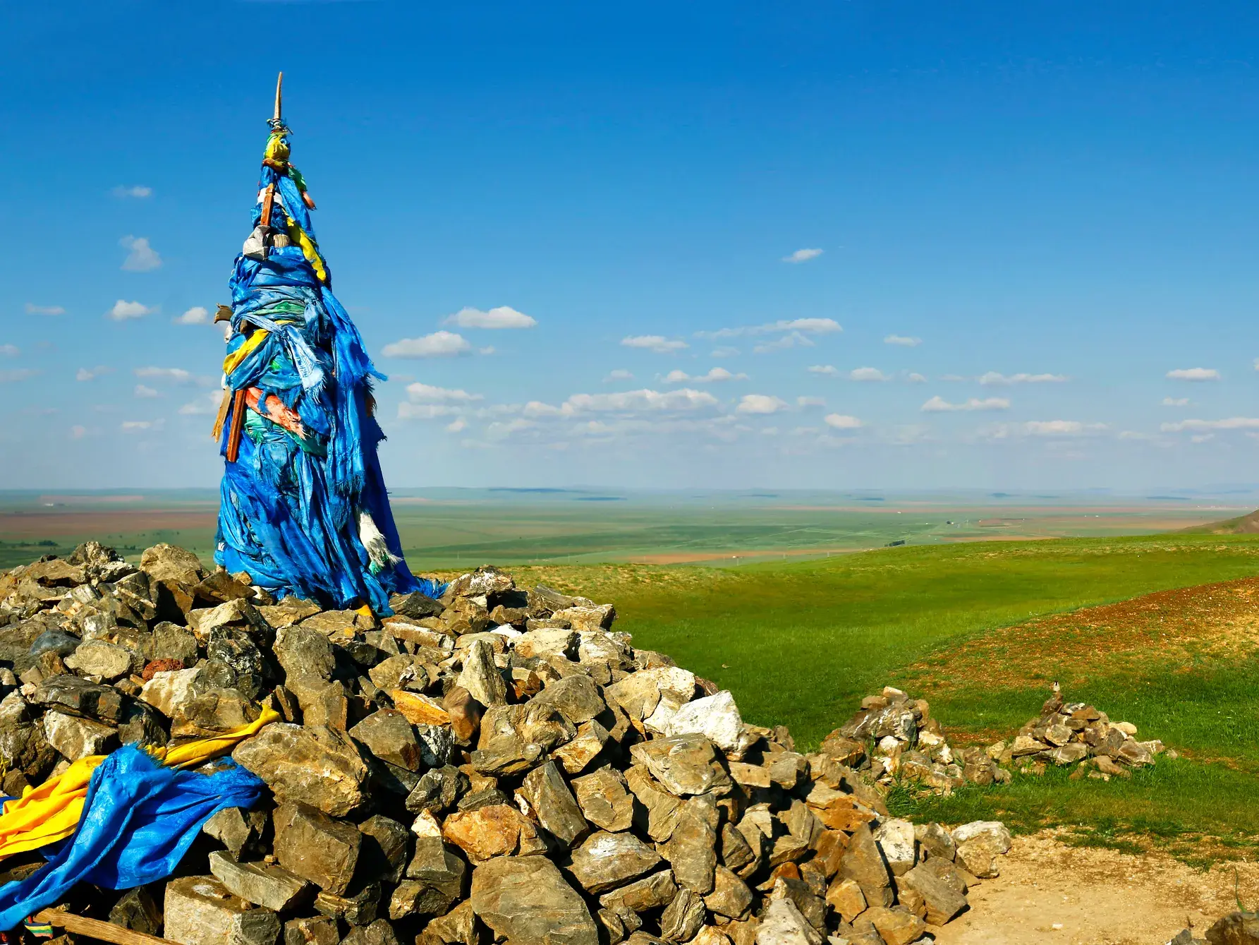 Ein traditioneller Owoo, ein mit blauen Tüchern geschmückter Steinhügel, in der endlosen, grünen Steppenlandschaft der Mongolei.