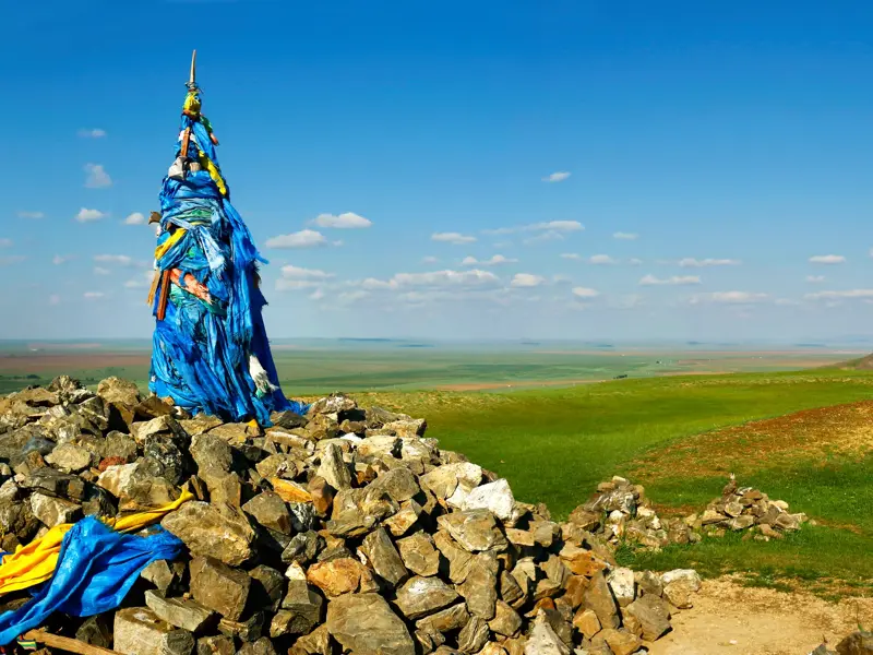 Ein traditioneller Owoo, ein mit blauen Tüchern geschmückter Steinhügel, in der endlosen, grünen Steppenlandschaft der Mongolei.