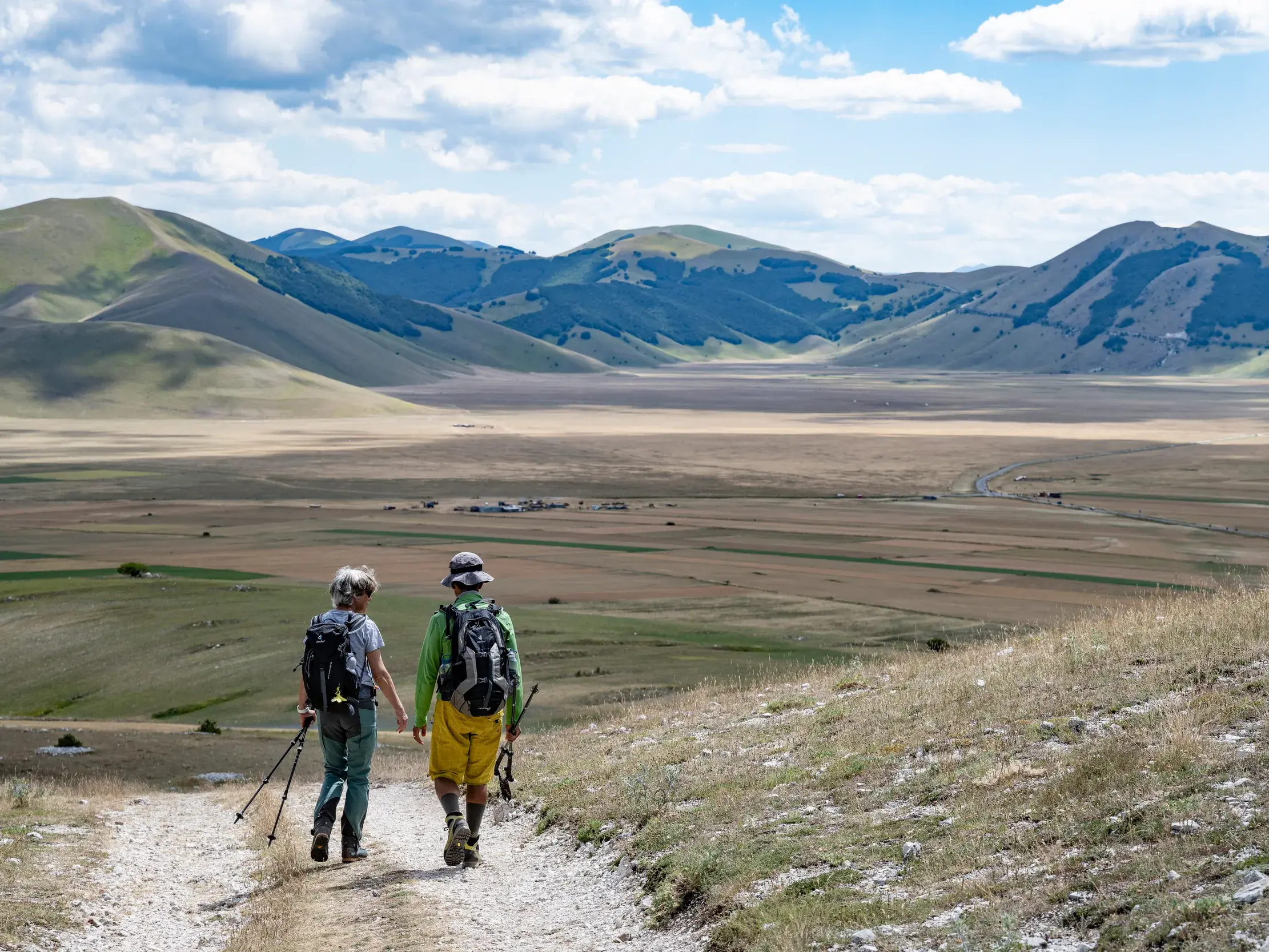 Zwei Wanderer gehen bergab auf einem Pfad mit Panoramablick über die Hochebene Piano Grande bei Castelluccio di Norcia.