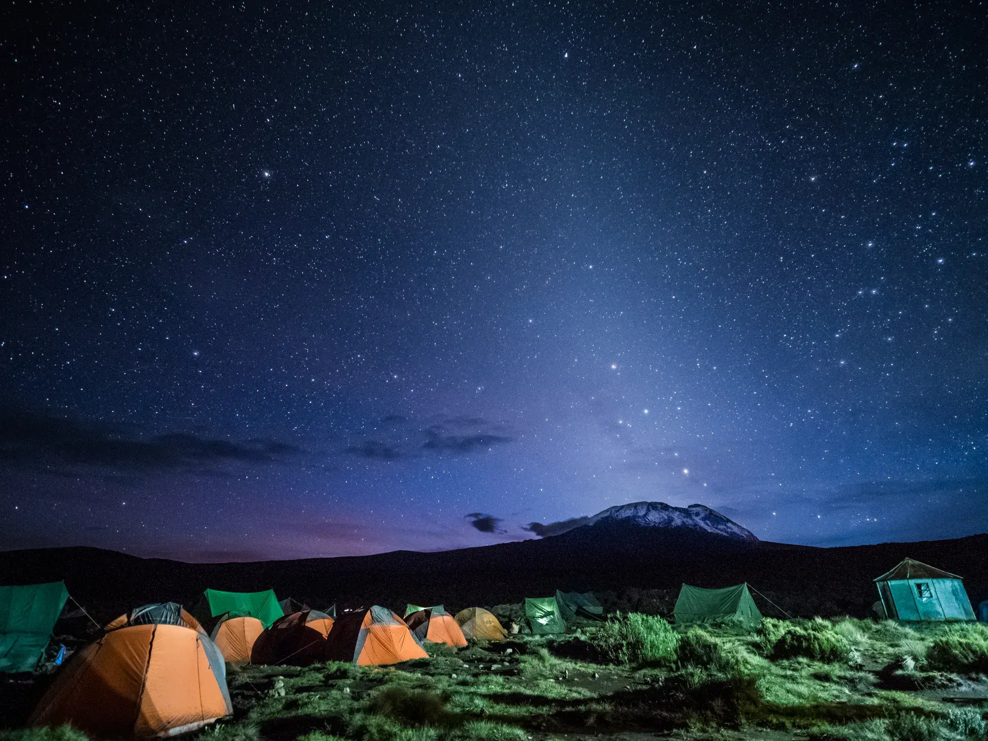 Nächtliches Zeltlager einer Trekking-Gruppe am Kilimanjaro mit Blick auf den schneebedeckten Gipfel unter klarem Sternenhimmel.
