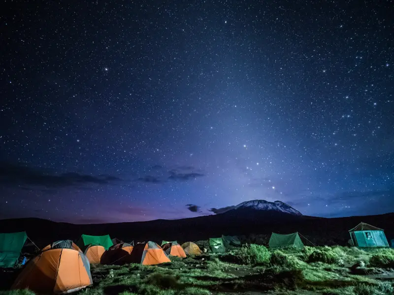 Nächtliches Zeltlager einer Trekking-Gruppe am Kilimanjaro mit Blick auf den schneebedeckten Gipfel unter klarem Sternenhimmel.