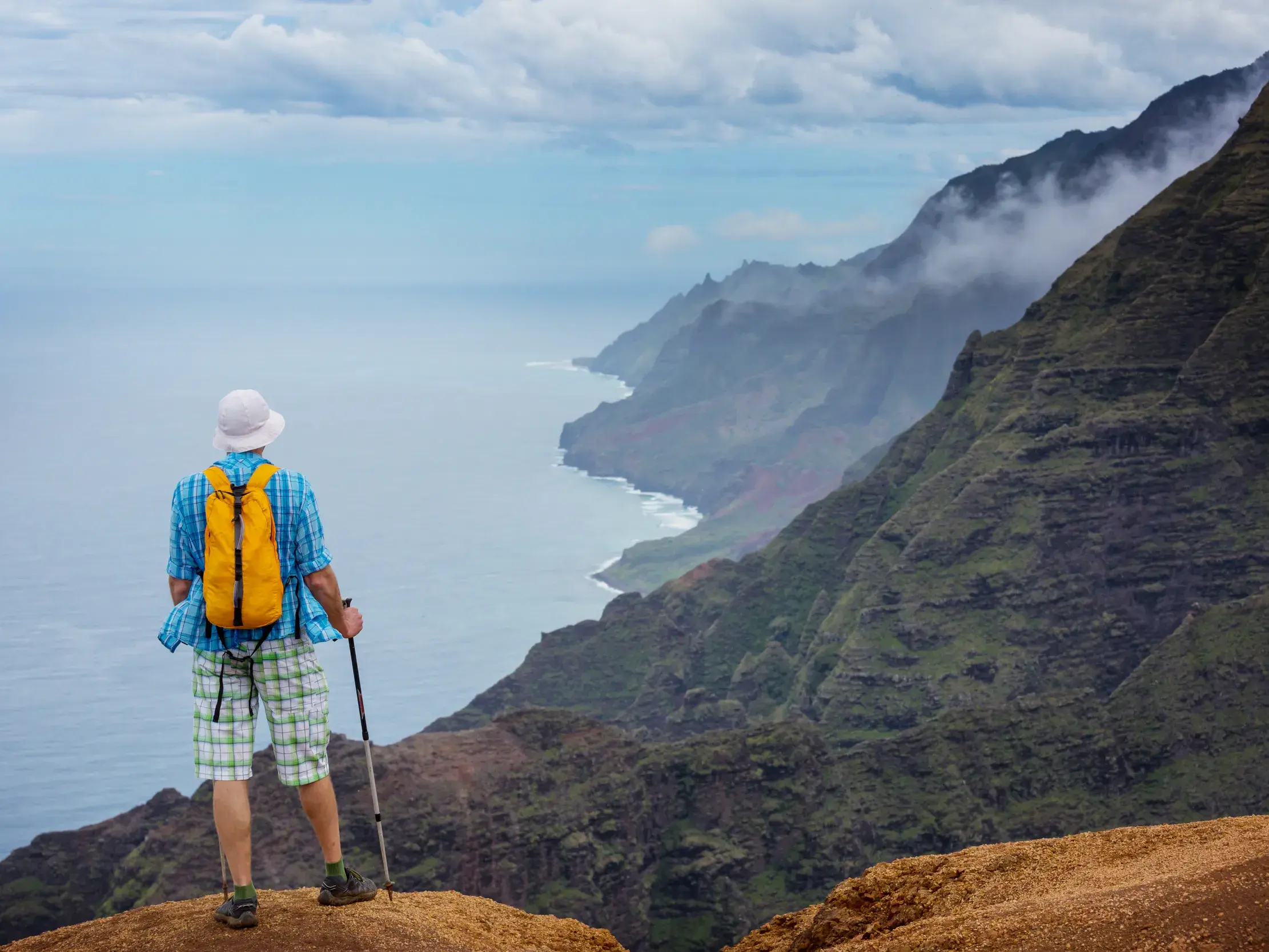 Ein Wanderer mit gelbem Rucksack blickt von den Nualolo Klippen auf die nebelverhangene, grüne Napali-Küste auf Kauai, Hawaii.