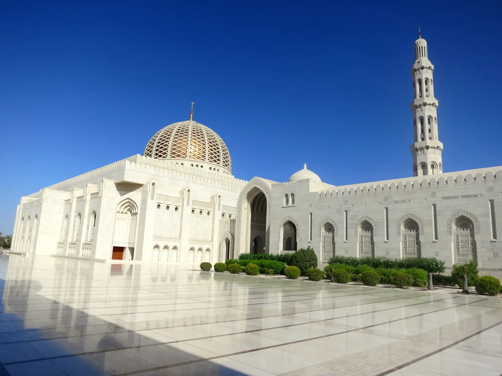 Die prächtige, weiße Sultan-Qabus-Moschee in Maskat mit ihrer goldenen Kuppel und dem hohen Minarett unter blauem Himmel.