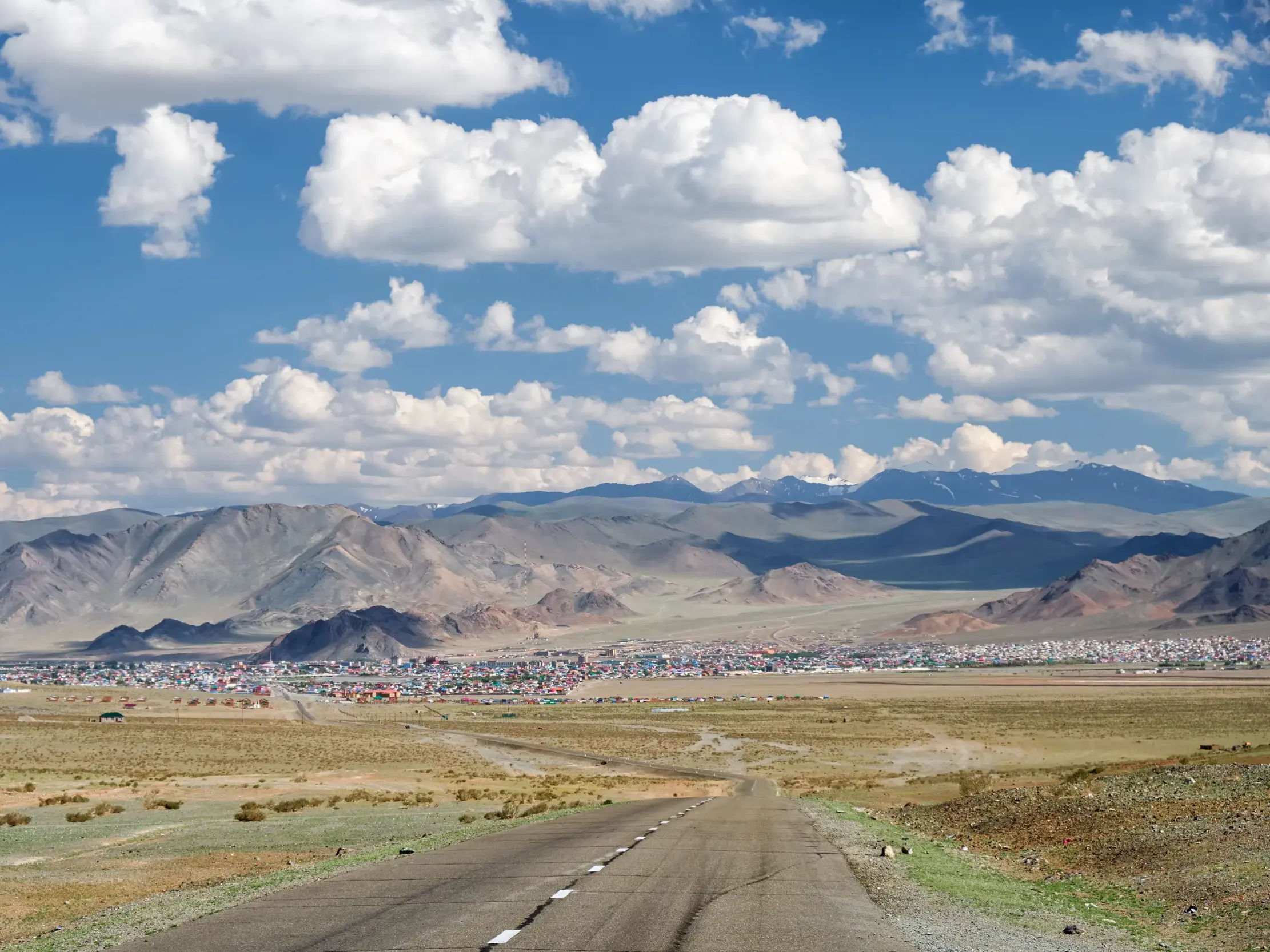 Eine Straße führt durch die weite Steppe der Westmongolei auf die Stadt Ölgii zu, im Hintergrund das Altai-Gebirge.