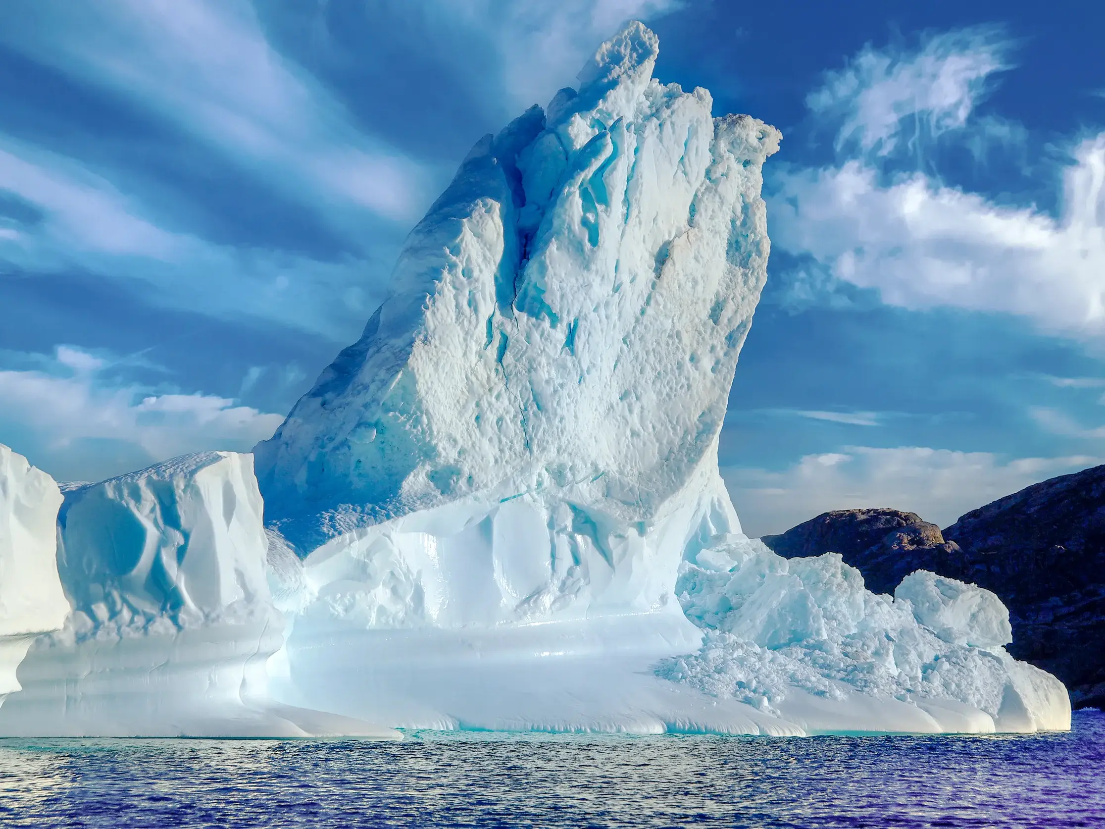 Ein majestätischer, spitzer Eisberg treibt im tiefblauen Polarmeer vor der felsigen Küste Ostgrönlands bei Ammassalik.