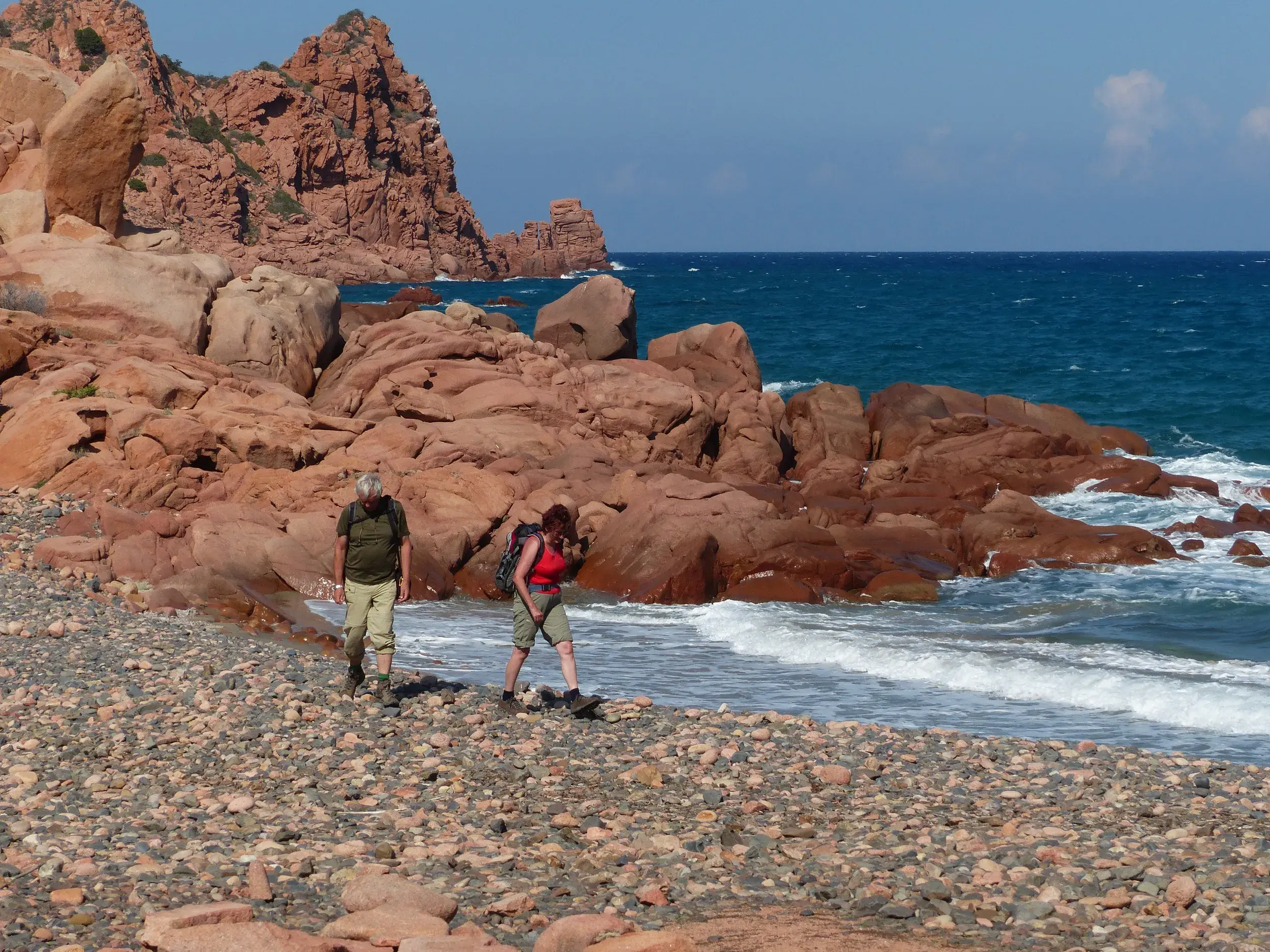 Zwei Wanderer mit Rucksäcken erkunden einen Steinstrand an der zerklüfteten, roten Felsenküste Sardiniens am Meer.
