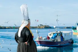 Frau in bretonischer Tracht mit hoher Spitzenhaube steht am Hafen und blickt auf ein Fischerboot in der Bretagne.