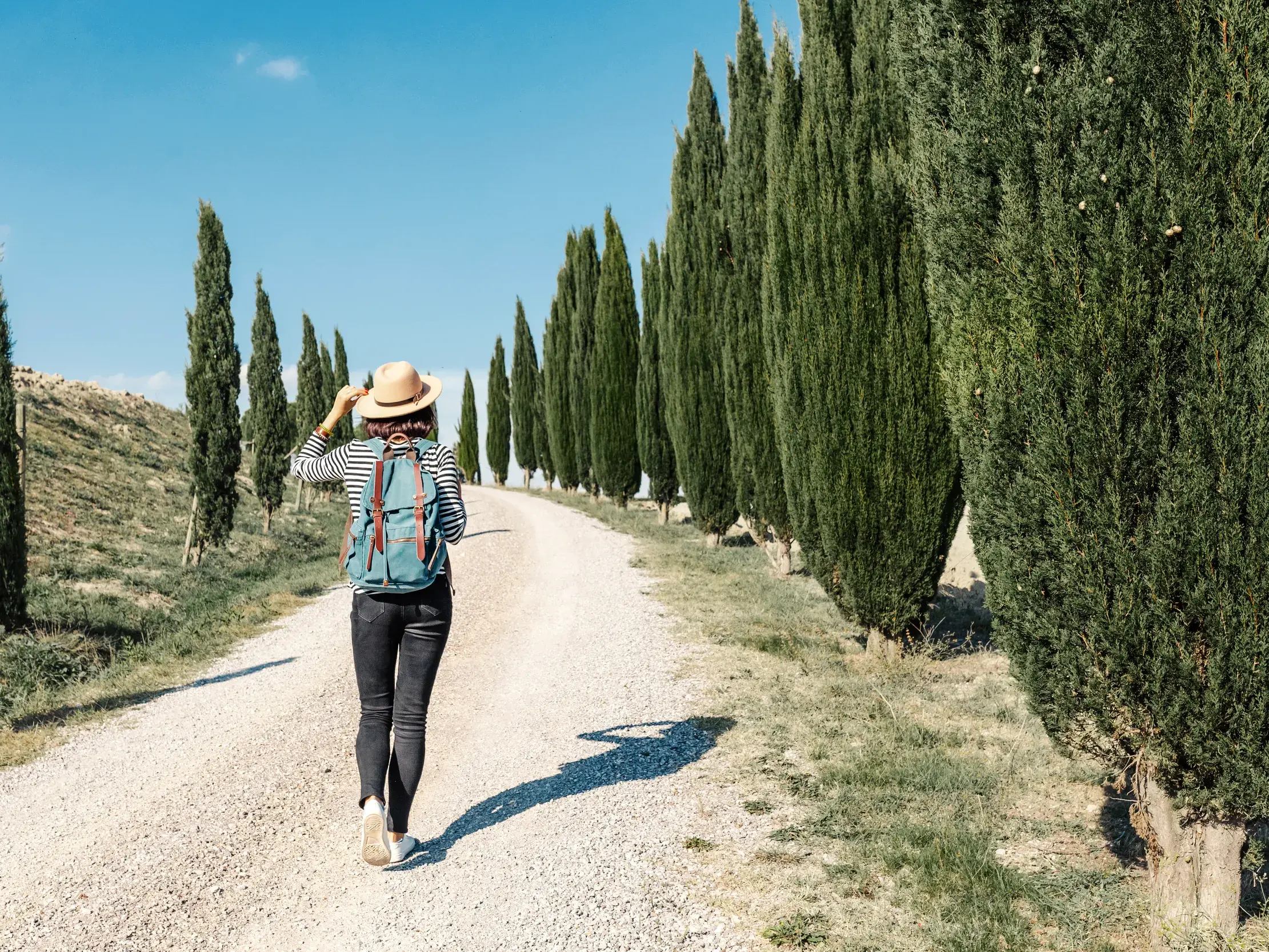 Eine Alleinreisende erkundet auf einer Wanderung eine Zypressenallee in der sonnigen Hügellandschaft der Toskana.