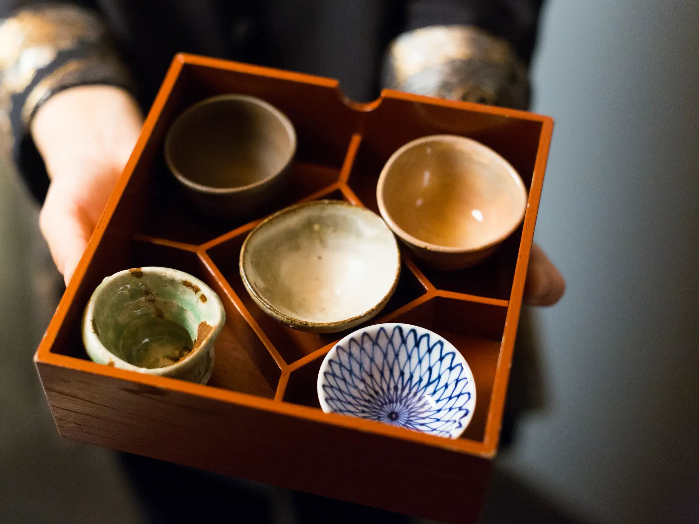 Hände präsentieren eine Auswahl japanischer Keramikschalen in einer Holzkiste für eine Sake-Verkostung während einer Studienreise.