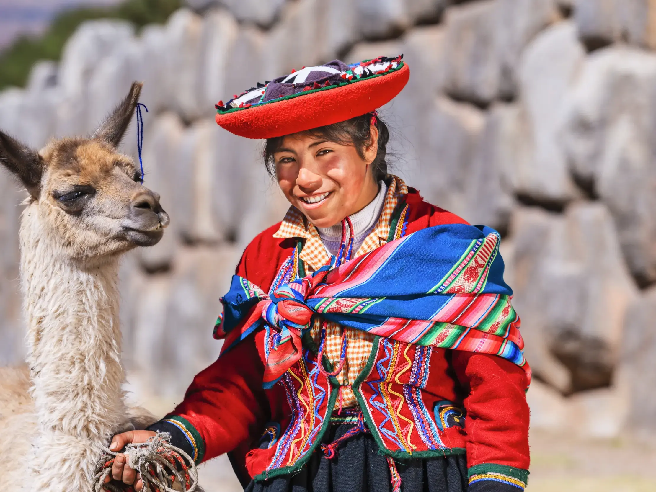 Eine lächelnde Peruanerin in traditioneller Tracht posiert mit ihrem Lama vor den Inka-Mauern von Sacsayhuamán bei Cusco.