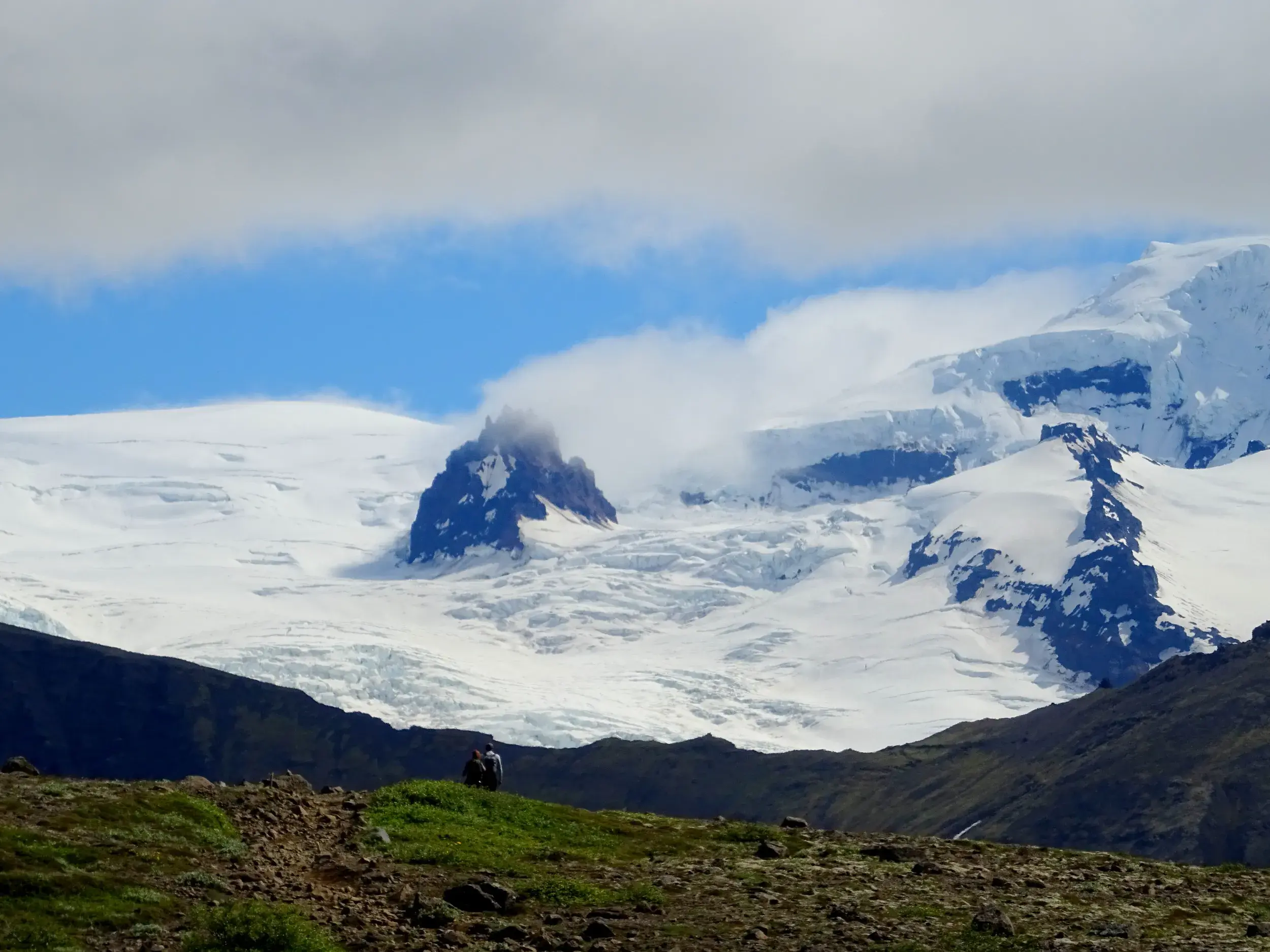 Zwei Wanderer pausieren auf einem grünen Hügel und blicken auf die gewaltige Gletscherlandschaft des Vatnajökull in Island.