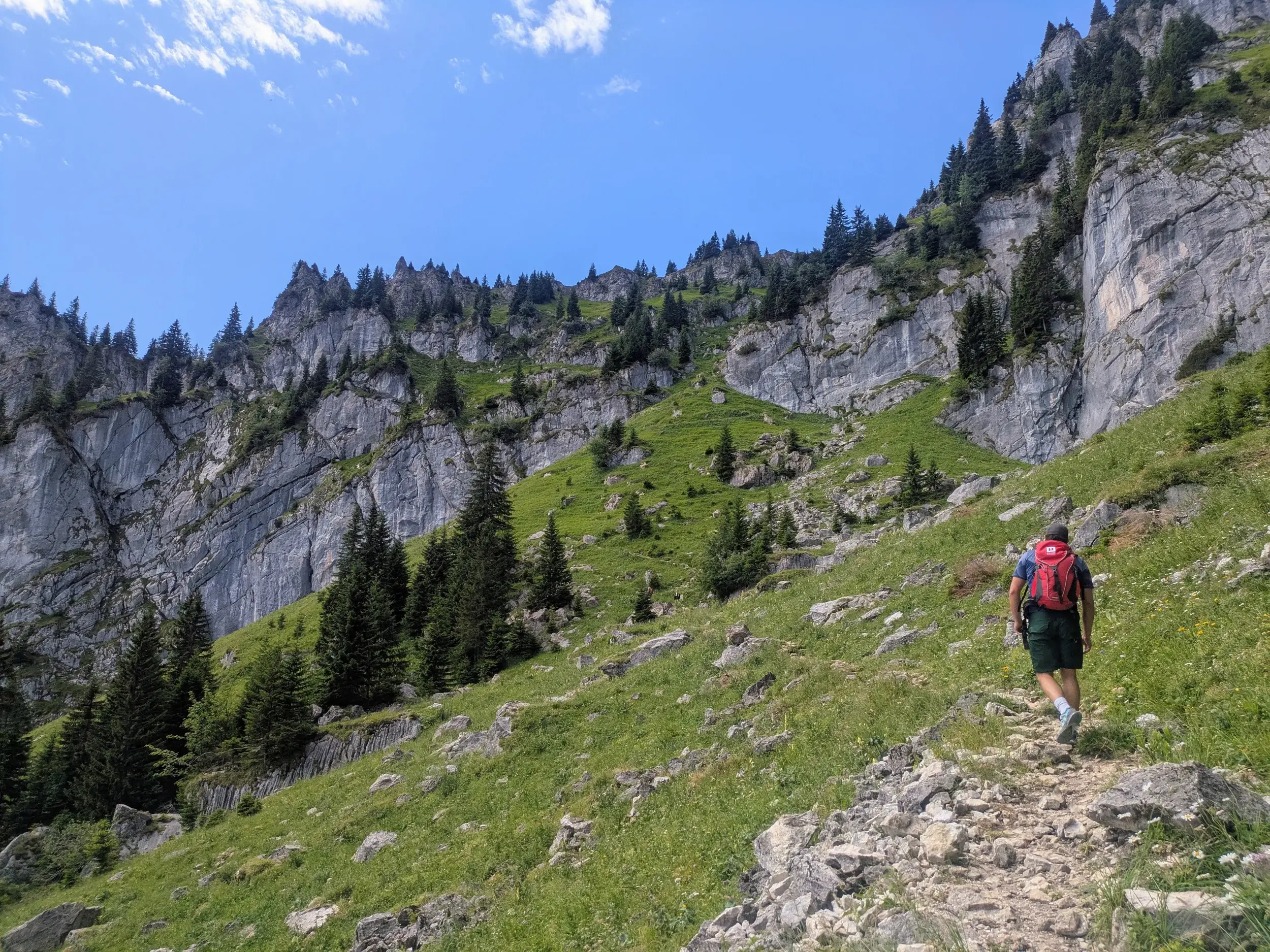 Ein Wanderer mit rotem Rucksack auf einem steinigen Pfad entlang einer steilen Felswand in den Ammergauer Alpen.