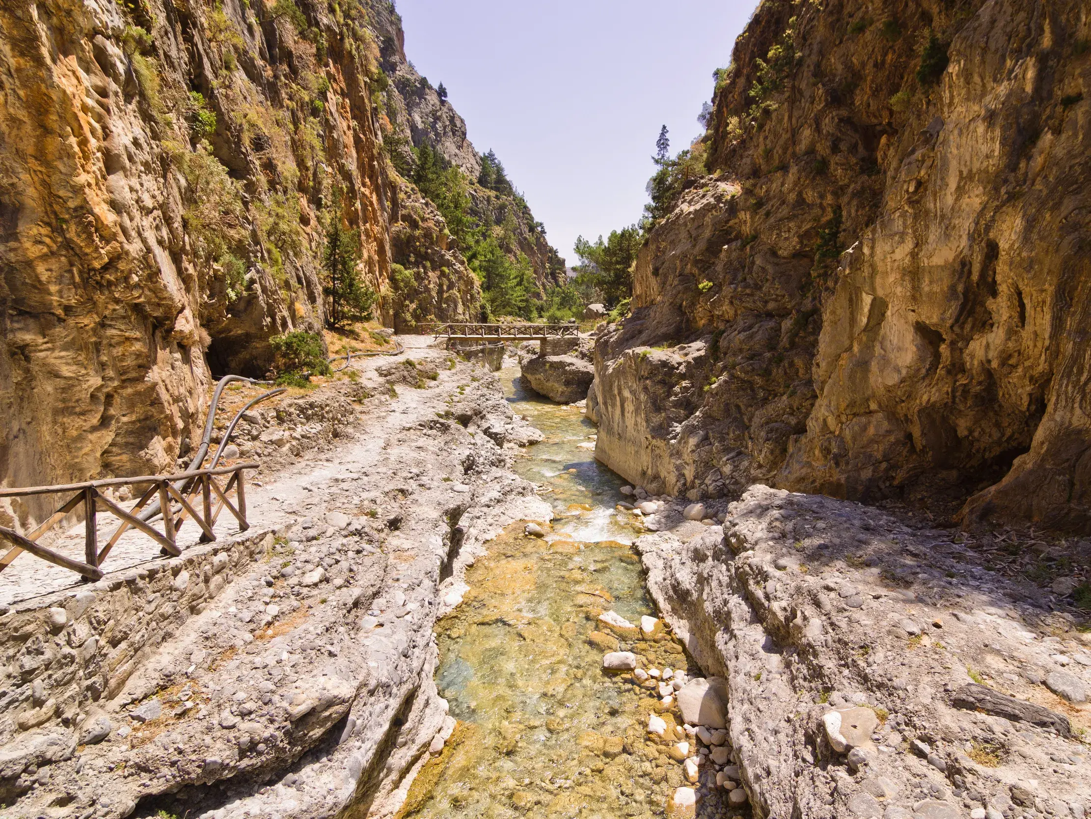 Ein steiniger Wanderpfad mit Holzgeländer verläuft neben einem klaren Bach durch die sonnenbeschienene Samaria-Schlucht auf Kreta.