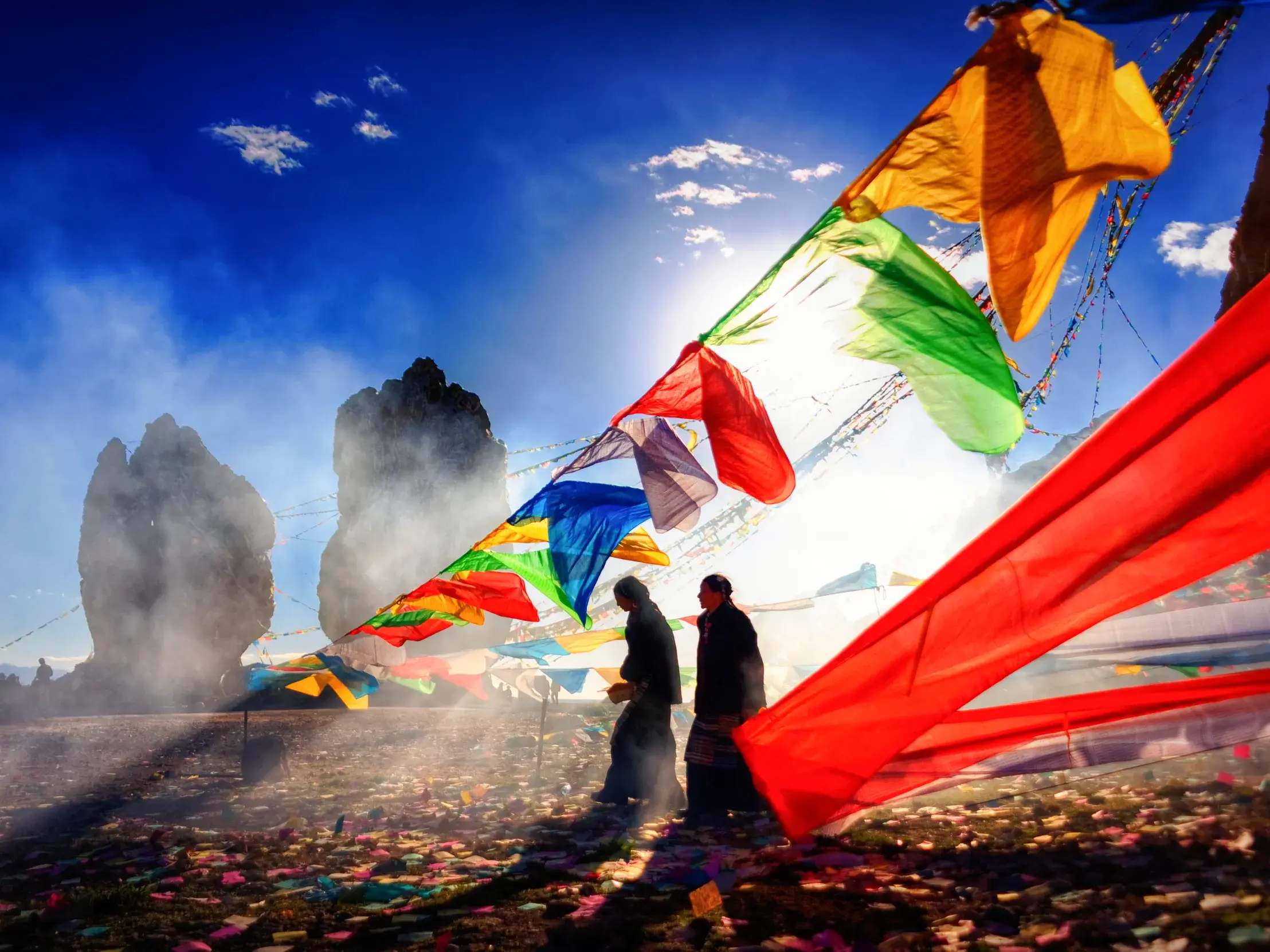 Zwei Pilger wandern unter bunten Gebetsfahnen im Gegenlicht der Sonne im Hochland von Tibet.