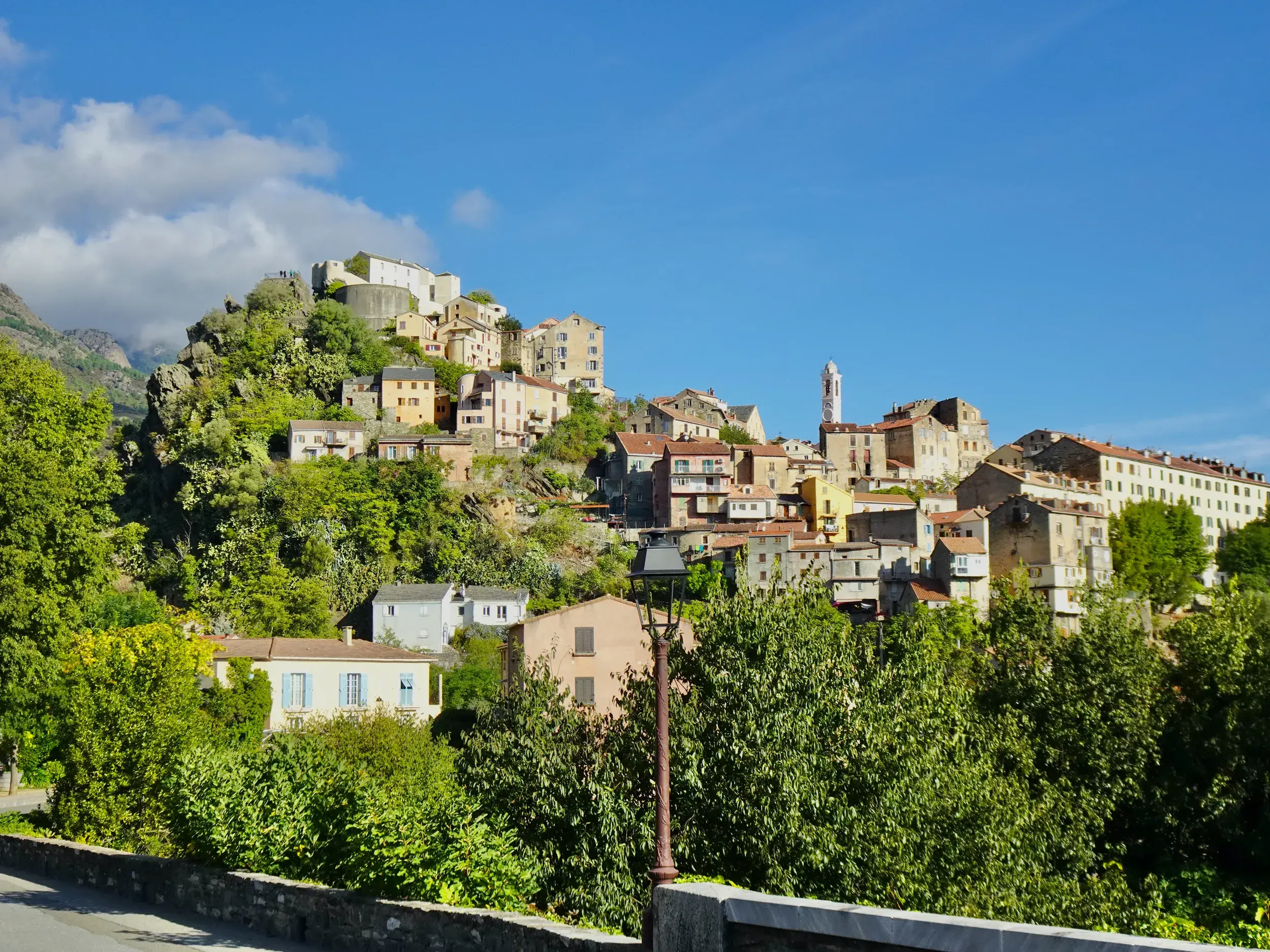 Die historische Altstadt von Corte auf Korsika thront auf einem Felsen, umgeben von grüner Macchia unter blauem Himmel.