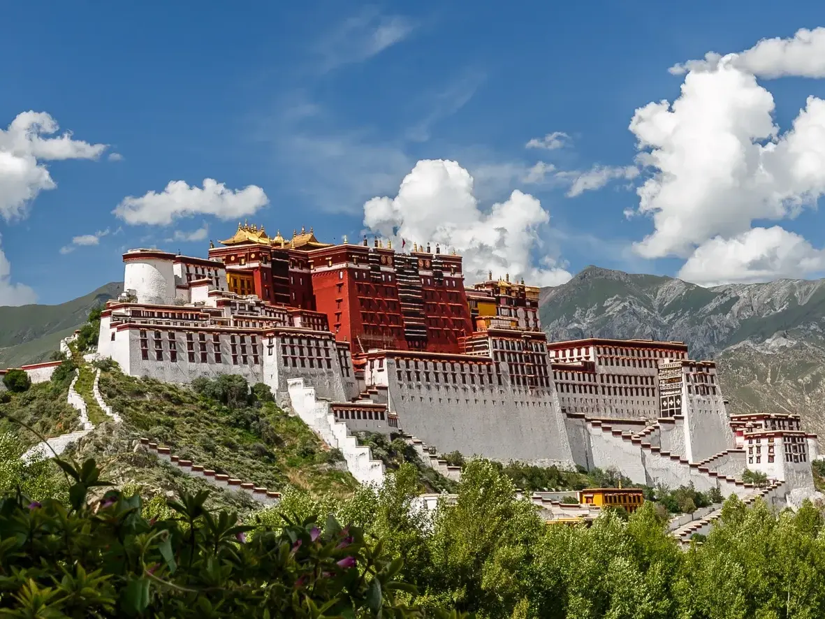 Der eindrucksvolle Potala-Palast in Lhasa thront auf dem Marpori Hügel vor einer Bergkette unter blauem Himmel.