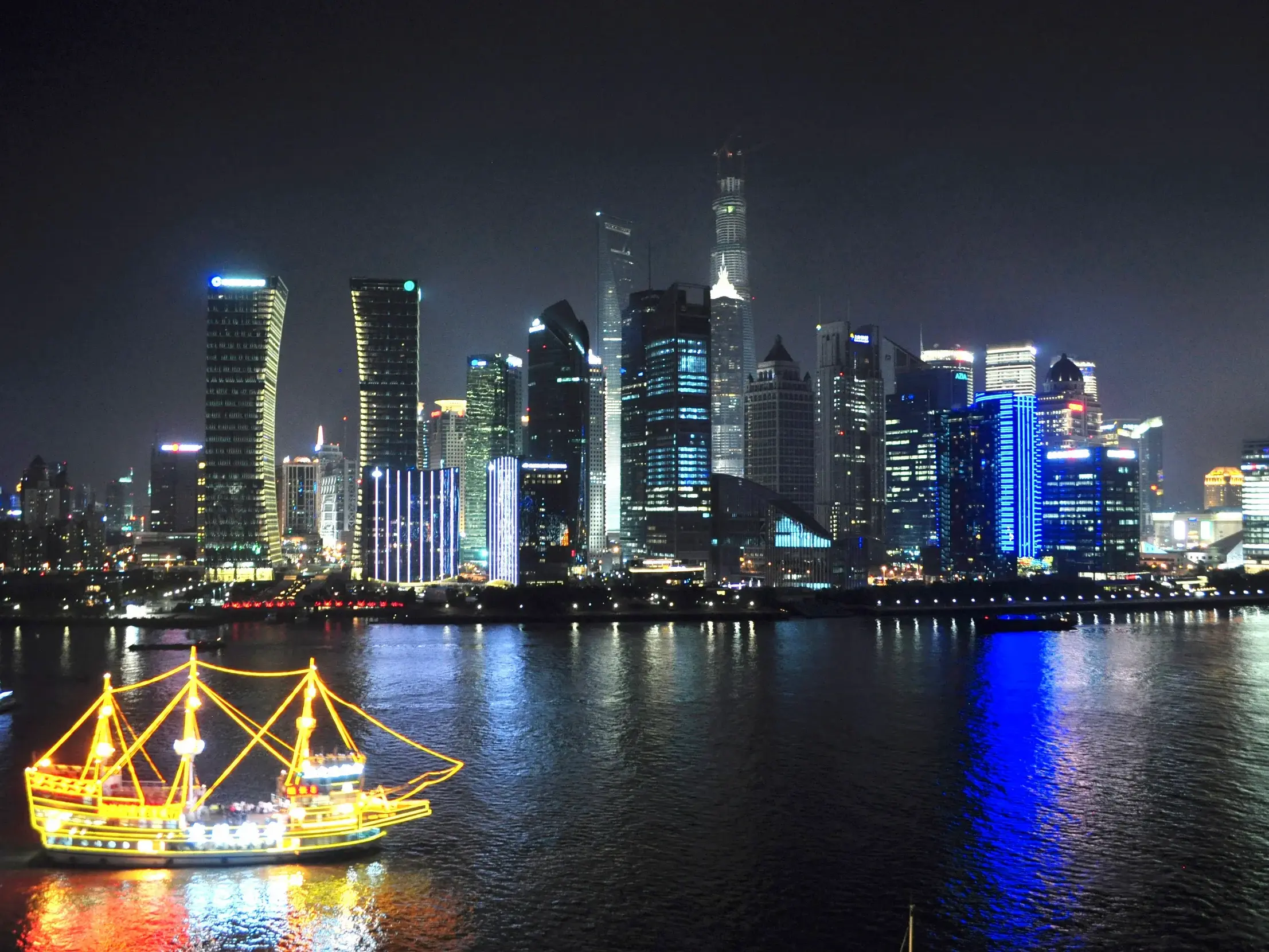 Hell erleuchtetes Ausflugsschiff auf dem Huangpu-Fluss vor der futuristischen Skyline von Pudong in Shanghai bei Nacht.