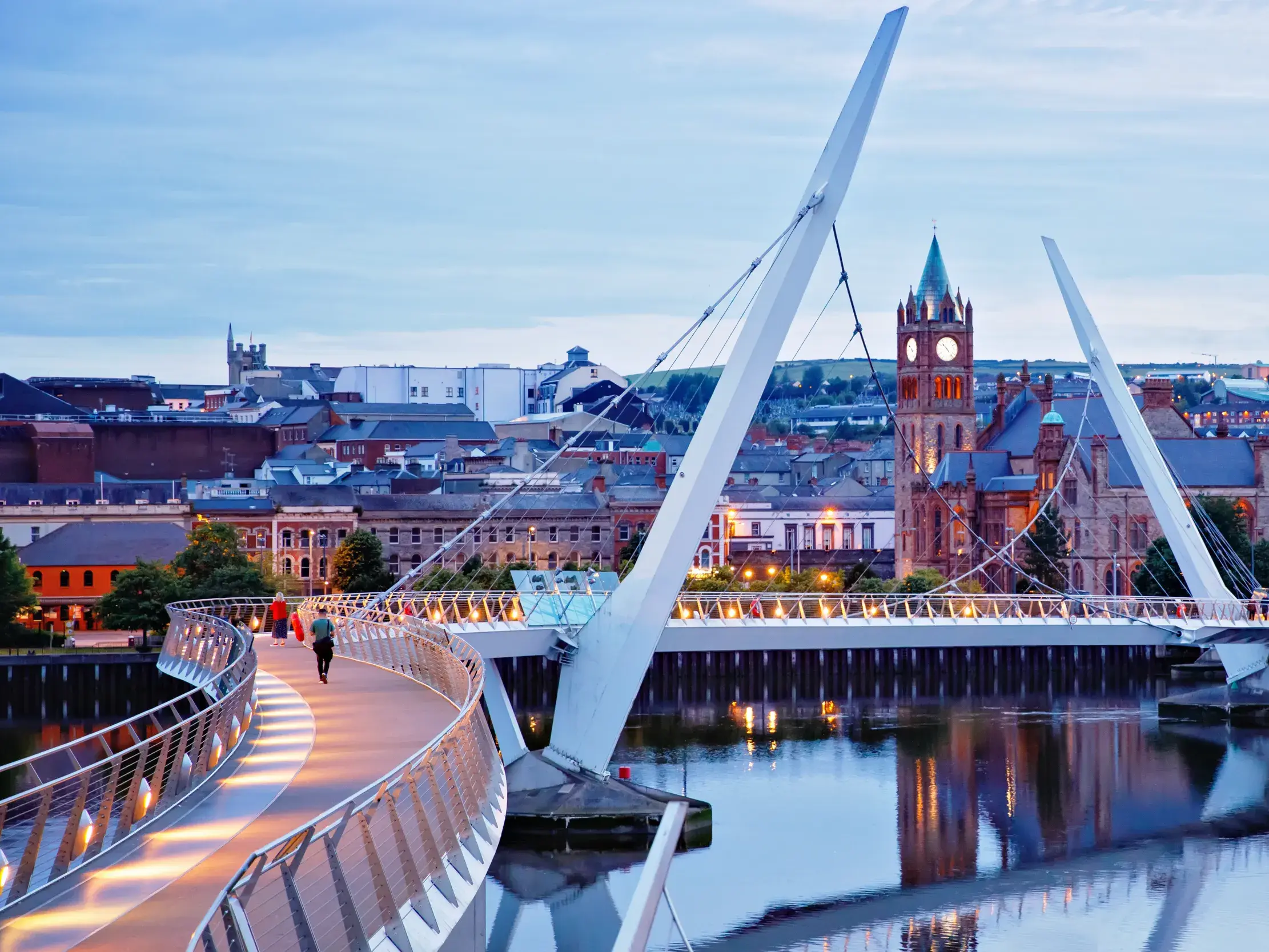 Fußgänger auf der beleuchteten Peace Bridge in Derry bei Dämmerung mit Blick auf die historische Guildhall am Fluss Foyle.