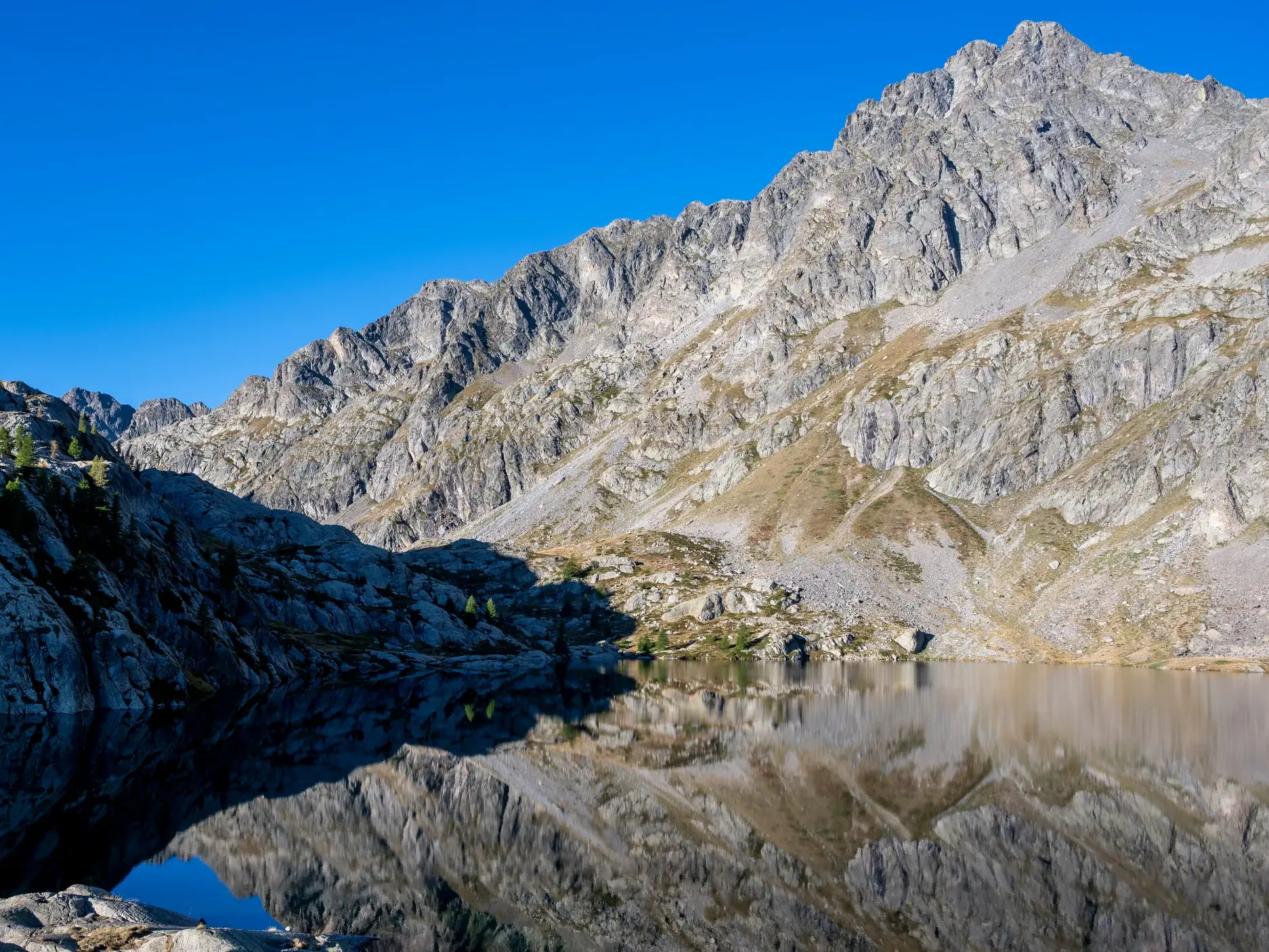 Ruhiger Bergsee bei einer Wanderung in den Seealpen, in dem sich die felsigen Gipfel unter strahlend blauem Himmel spiegeln.