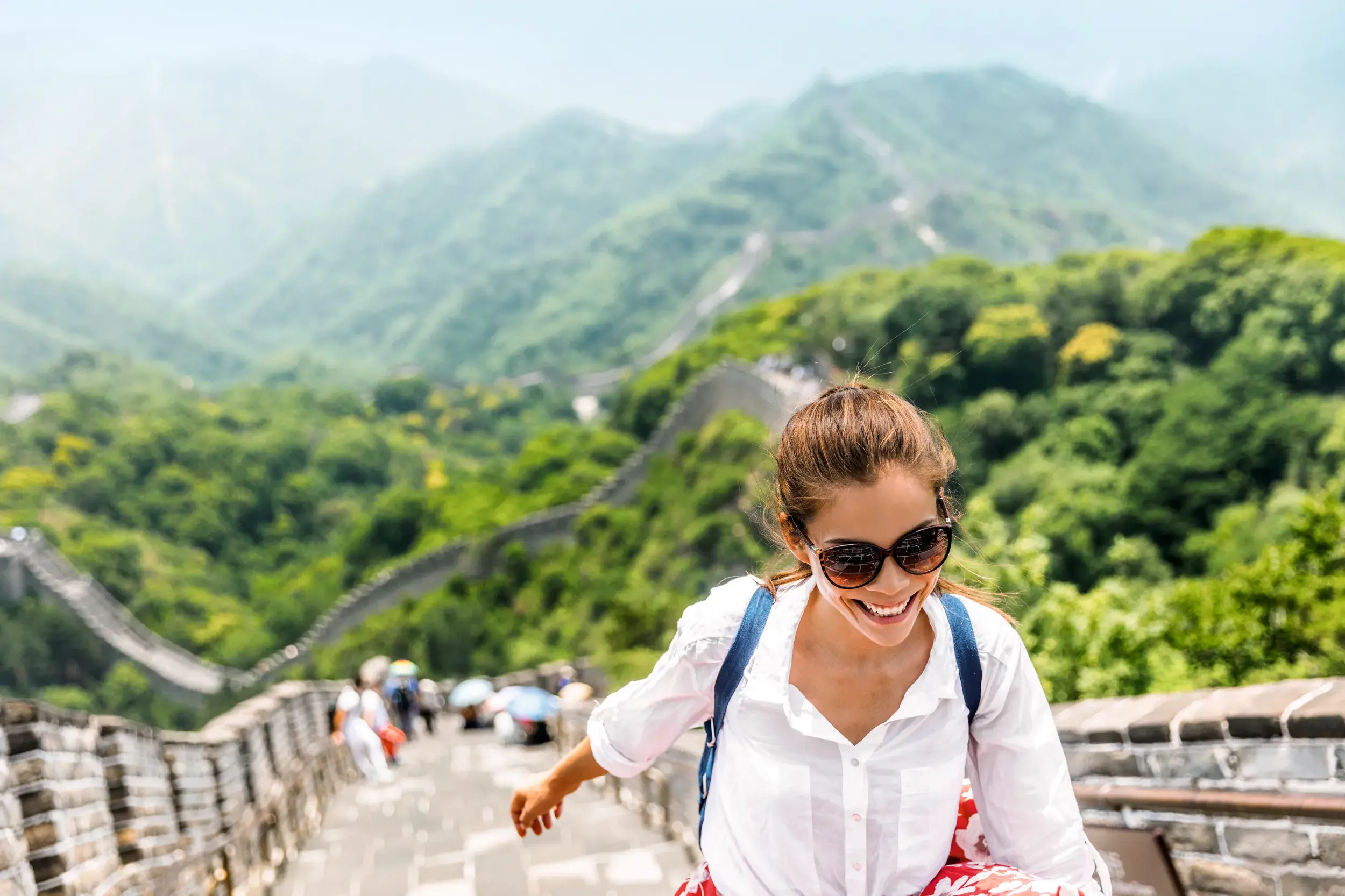 Fröhliche junge Frau mit Rucksack steigt Stufen auf der Großen Mauer in China hinab, die sich durch grüne Berge schlängelt.