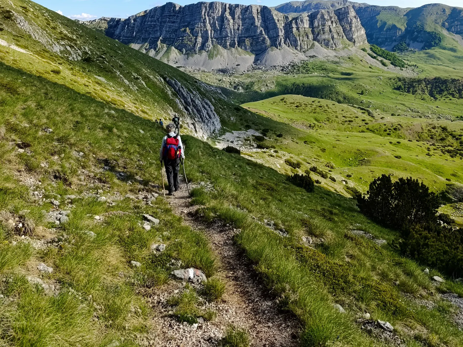 Eine Trekking-Gruppe wandert auf einem schmalen Pfad entlang eines sonnigen, grasbewachsenen Berghangs mit felsigen Gipfeln im Hintergrund.