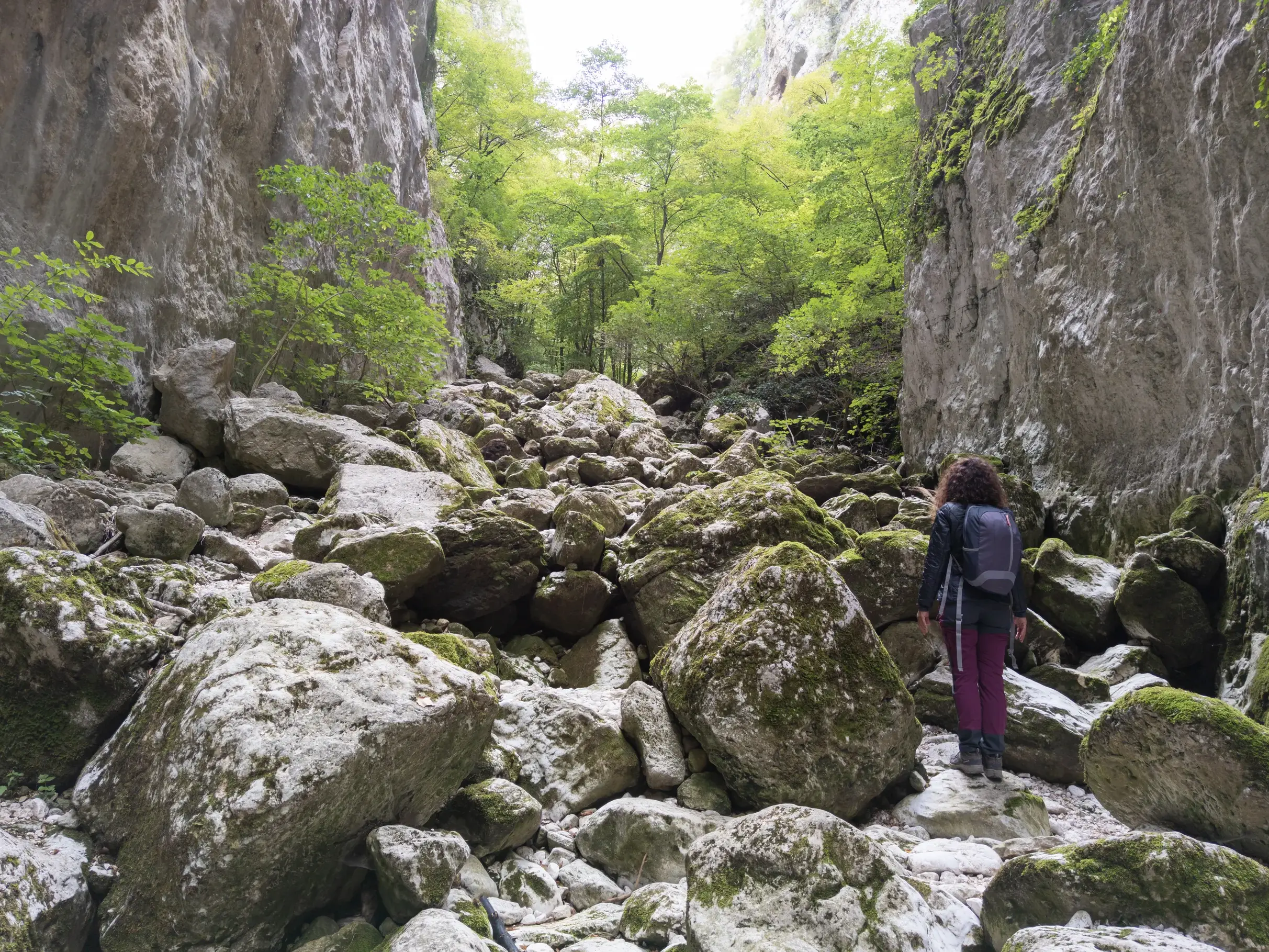 Eine Frau mit Rucksack wandert über große, moosbewachsene Felsen durch die enge Celano-Schlucht in den Abruzzen.