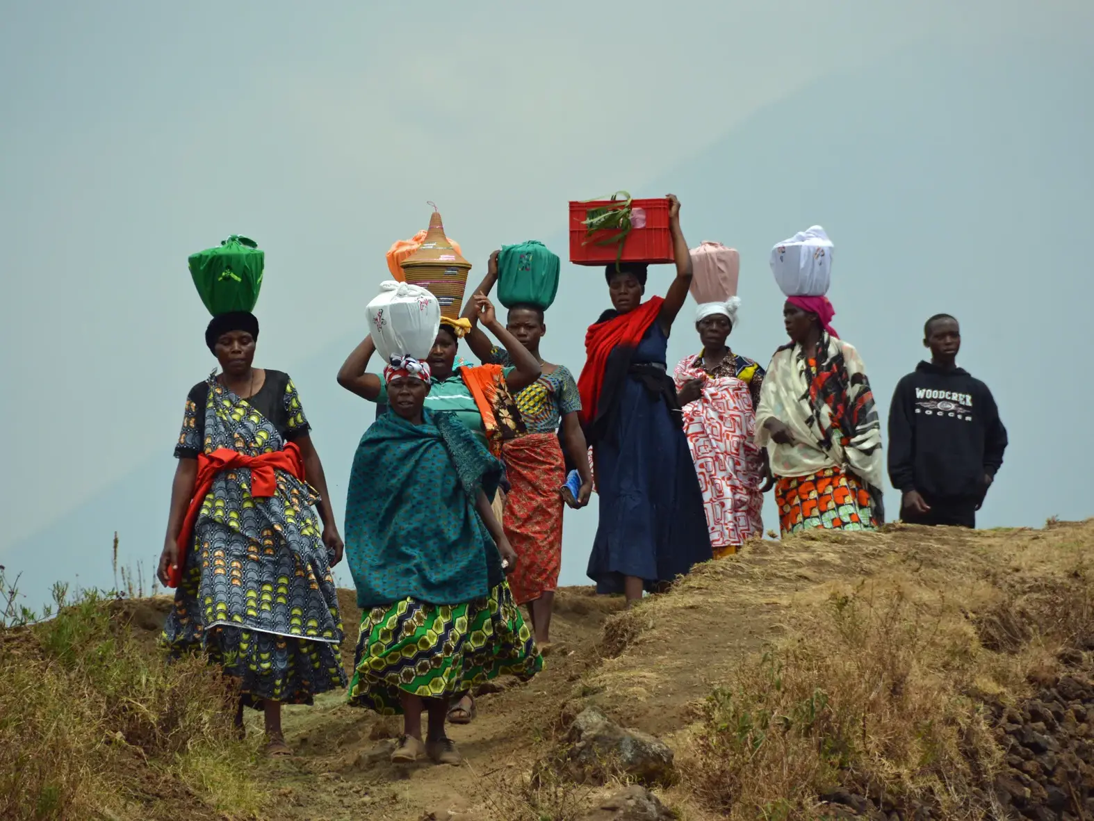 Gruppe einheimischer Frauen in bunten Kleidern trägt traditionell Körbe und Waren auf dem Kopf über einen erdigen Pfad in Ruanda.