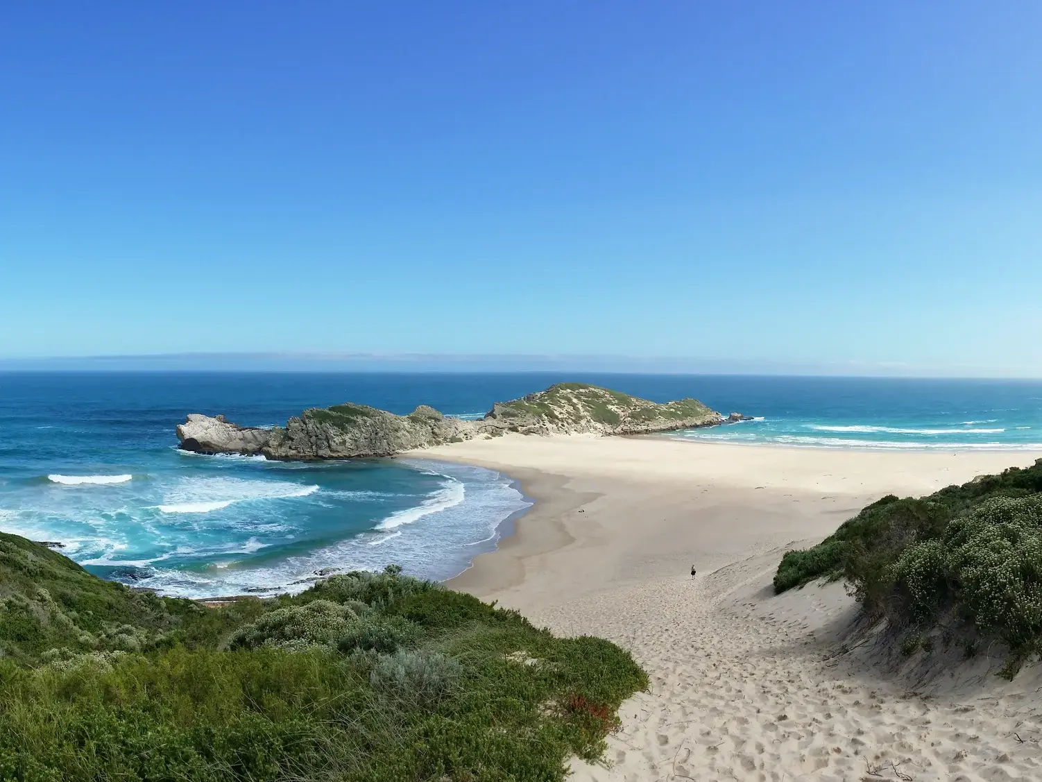 Blick von einer Düne auf einen weiten Sandstrand mit Felsen am türkisblauen Meer entlang der südafrikanischen Gartenroute.