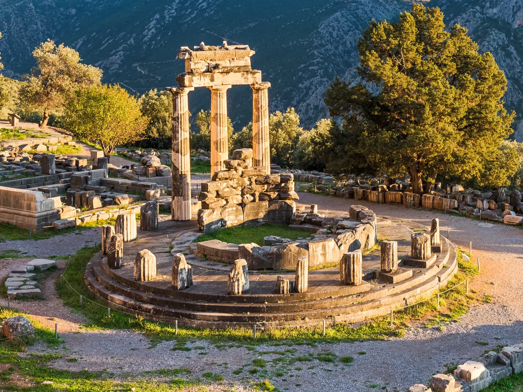 Die antiken Ruinen des Tholos-Heiligtums in Delphi, umgeben von grünen Bäumen vor der Bergkulisse des Parnassgebirges.