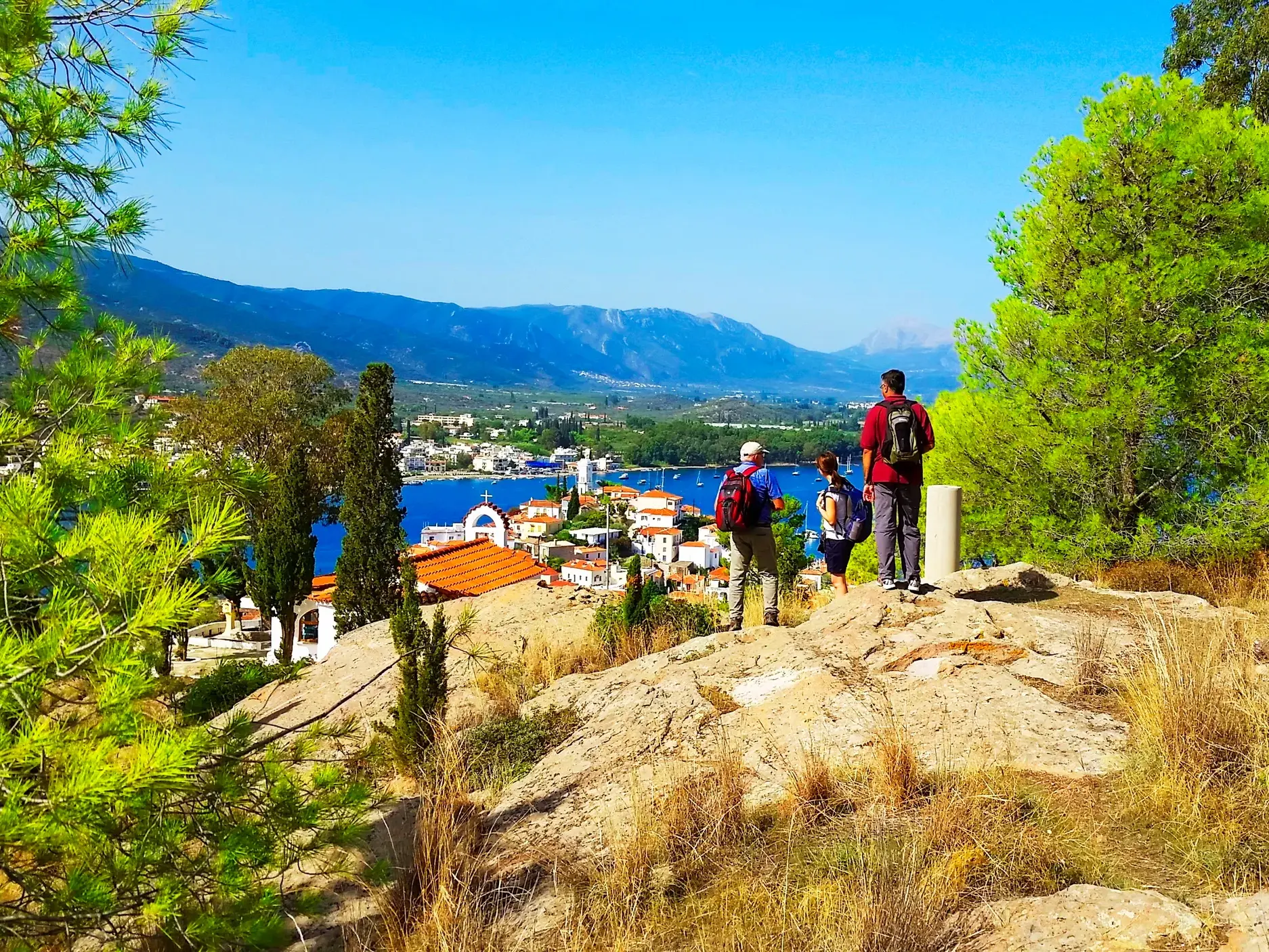 Drei Wanderer auf einer Anhöhe mit Panoramablick auf Poros-Stadt, umgeben von Pinien und dem blauen Saronischen Golf.