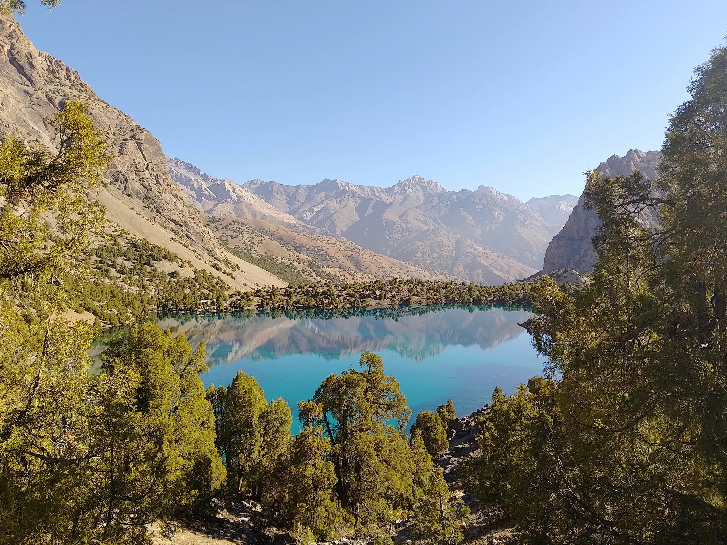 Blick von einem Wanderweg durch Wacholderbäume auf einen türkisen See, in dem sich die Berge des Fan-Gebirges spiegeln.