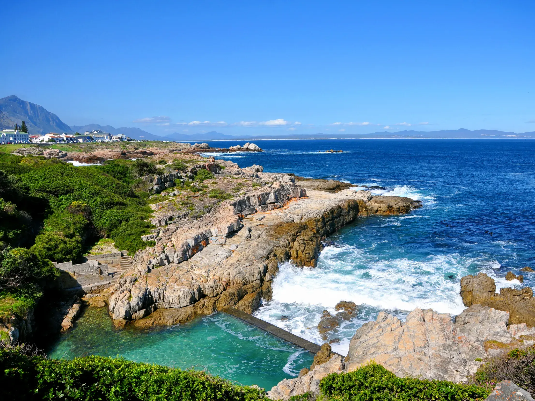 Ein türkisblaues Gezeitenbecken an der Felsenküste von Hermanus in Südafrika mit Blick auf den weiten, blauen Ozean.