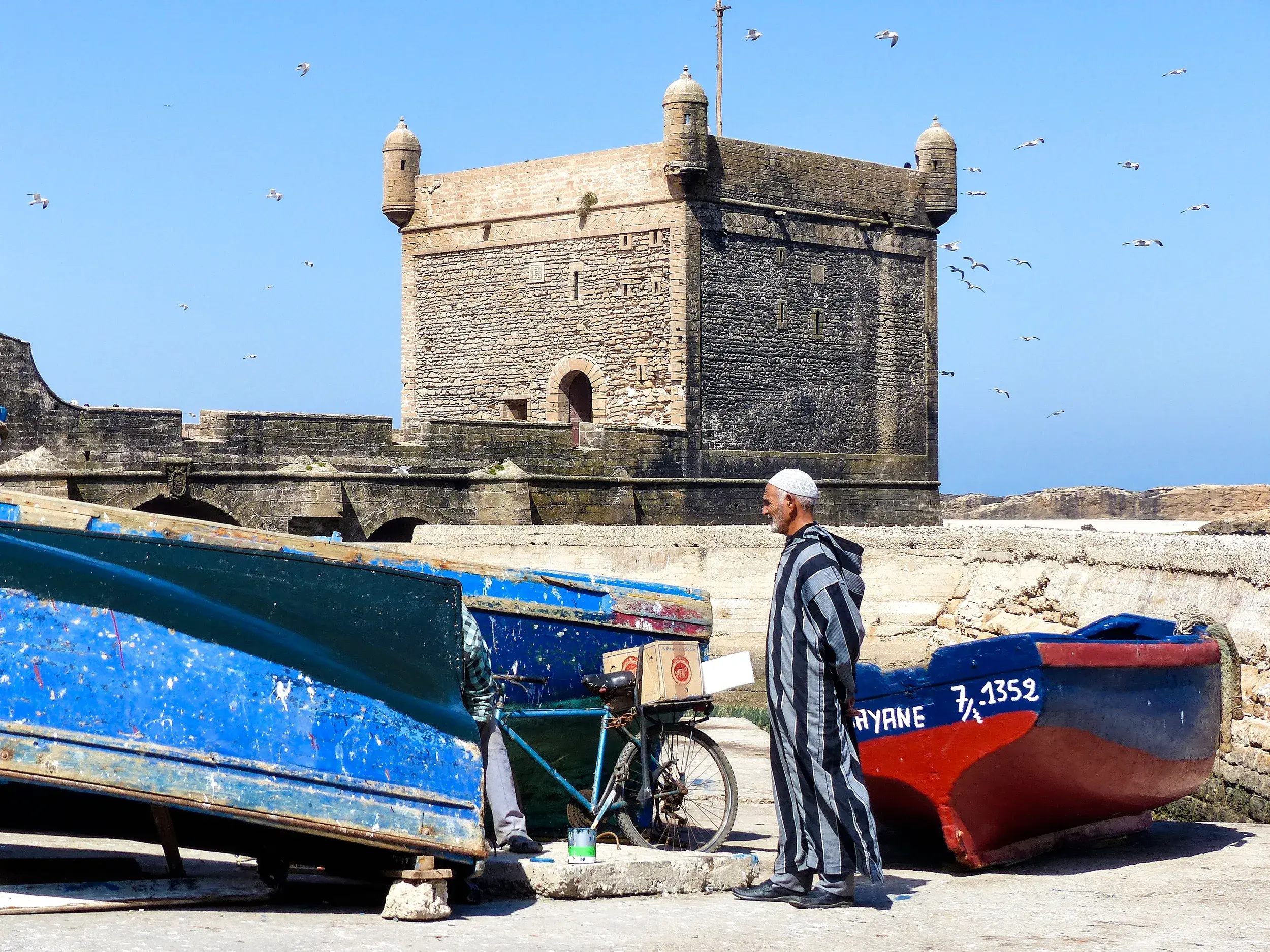 Ein Fischer in traditioneller Djellaba steht am Hafen von Essaouira vor der Festungsmauer und bunten Booten.