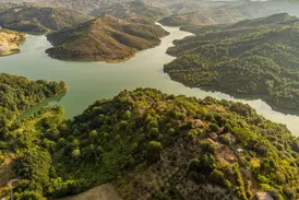 Luftaufnahme des türkisfarbenen Koman-Stausees in Albanien, der sich durch eine grüne, hügelige Fjordlandschaft schlängelt.