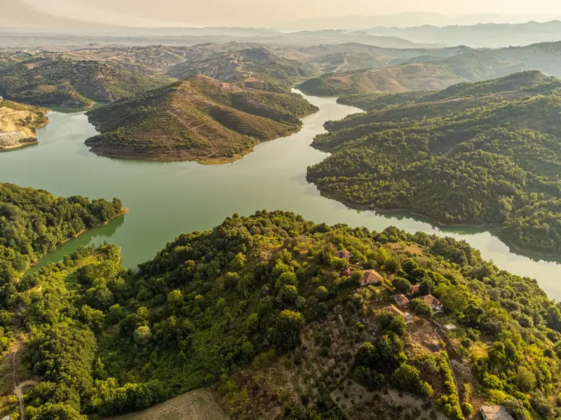 Luftaufnahme des türkisfarbenen Koman-Stausees in Albanien, der sich durch eine grüne, hügelige Fjordlandschaft schlängelt.