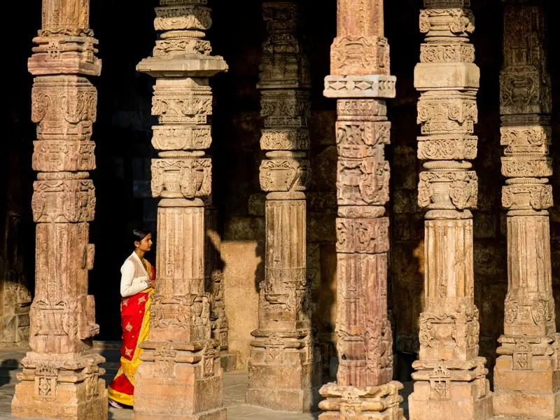 Eine Frau in rotem Sari bewundert die reich verzierten Sandsteinsäulen im Qutub Minar Komplex in Delhi.