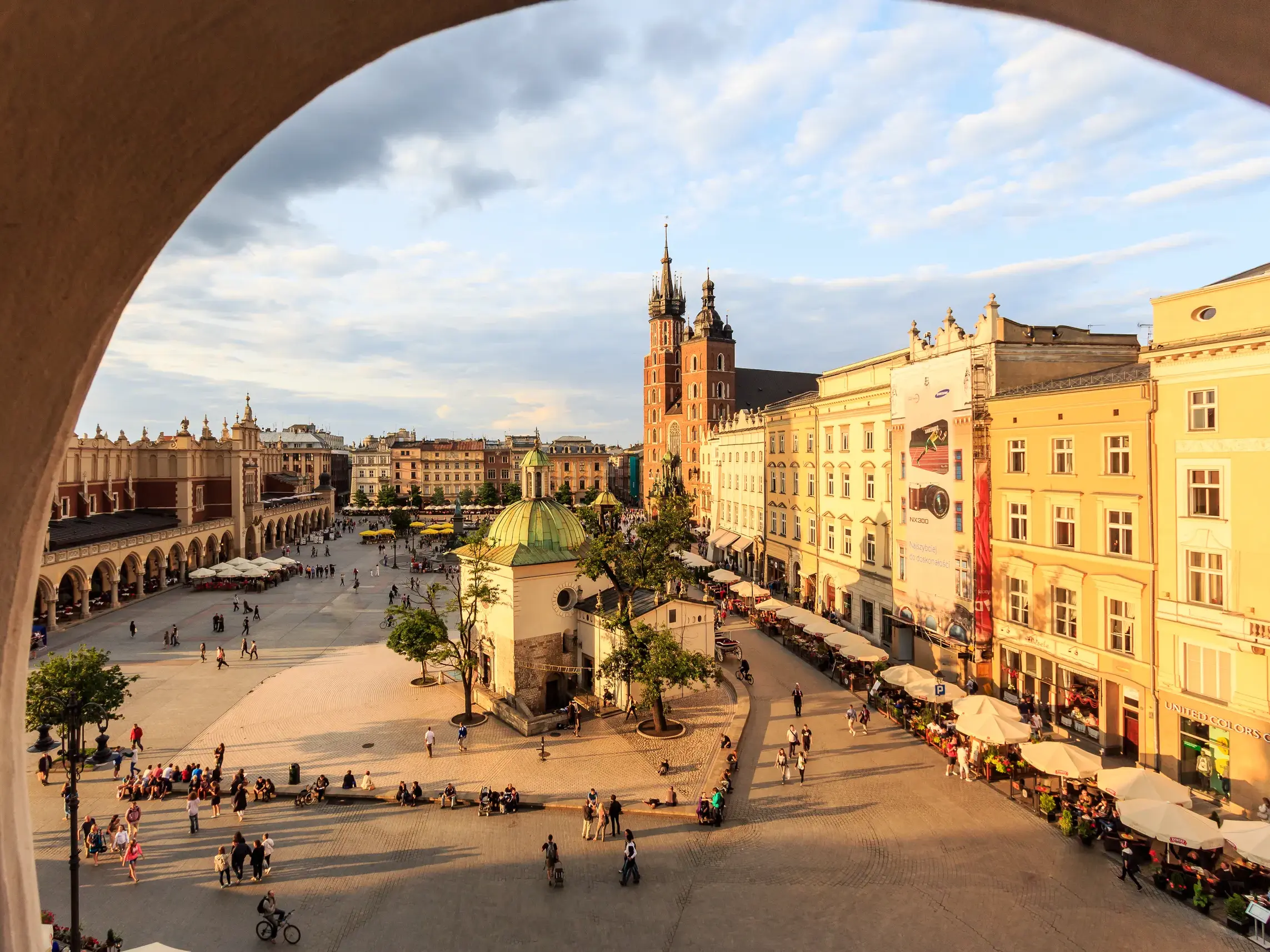 Blick durch einen Bogen auf den belebten Hauptring in Krakau mit den Tuchhallen und der Marienkirche in der Abendsonne.