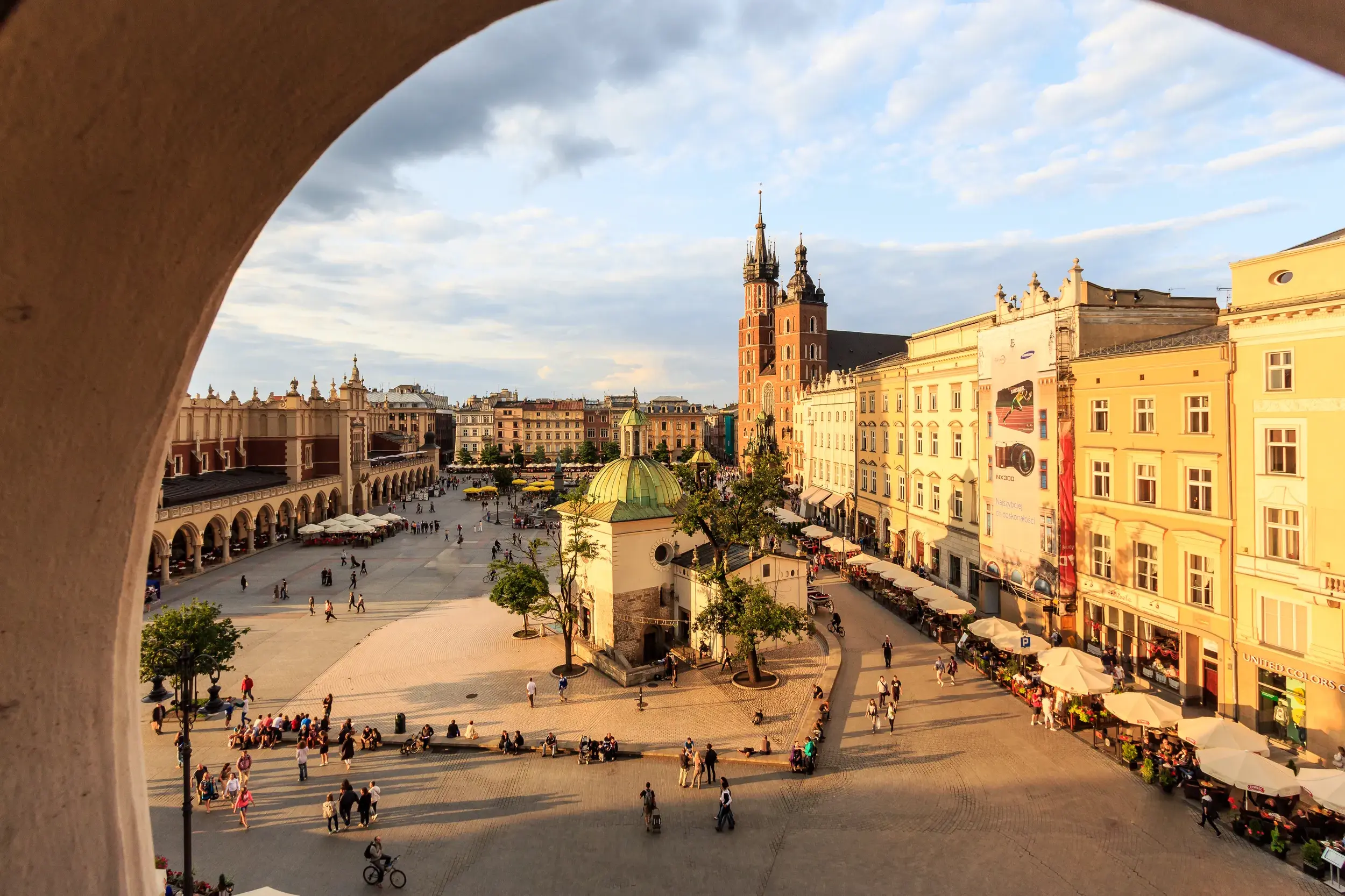 Blick durch einen Bogen auf den belebten Hauptring in Krakau mit den Tuchhallen und der Marienkirche in der Abendsonne.